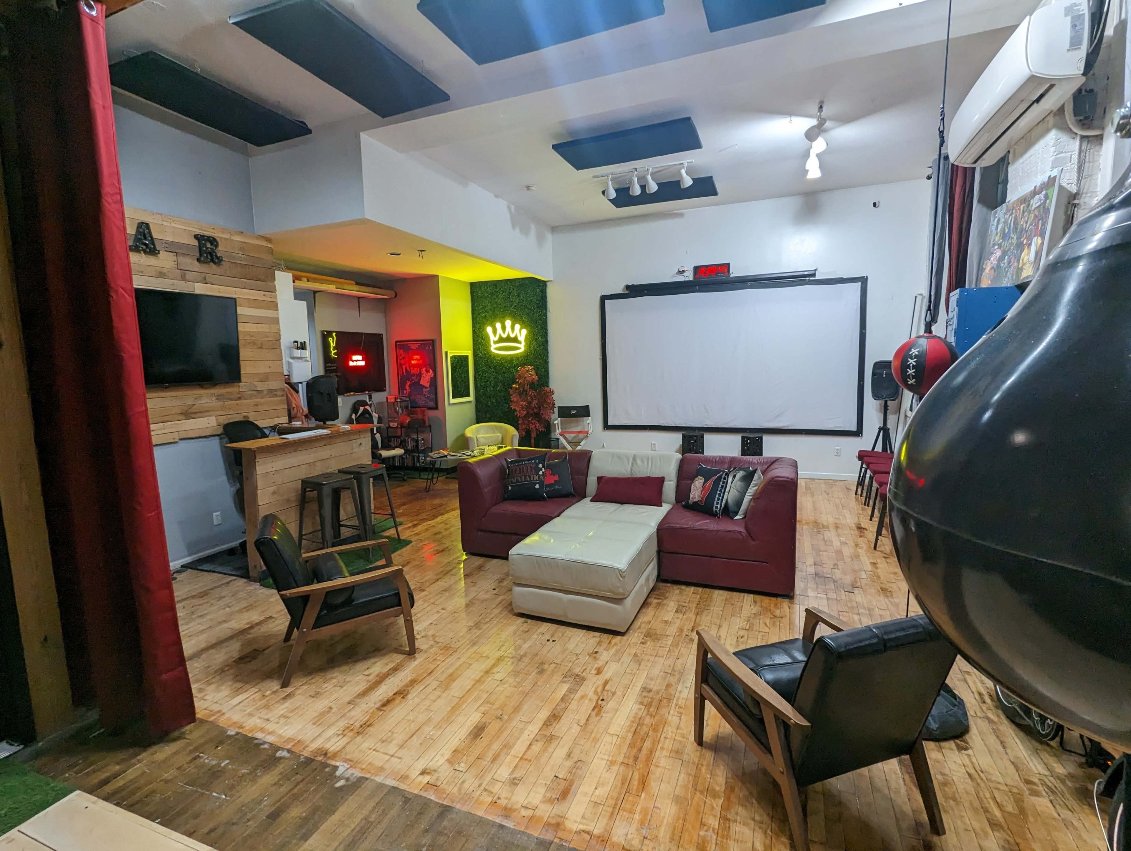 A spacious lounge area with a red couch, a large projection screen, wooden flooring, and a bar on one side with various seating arrangements.