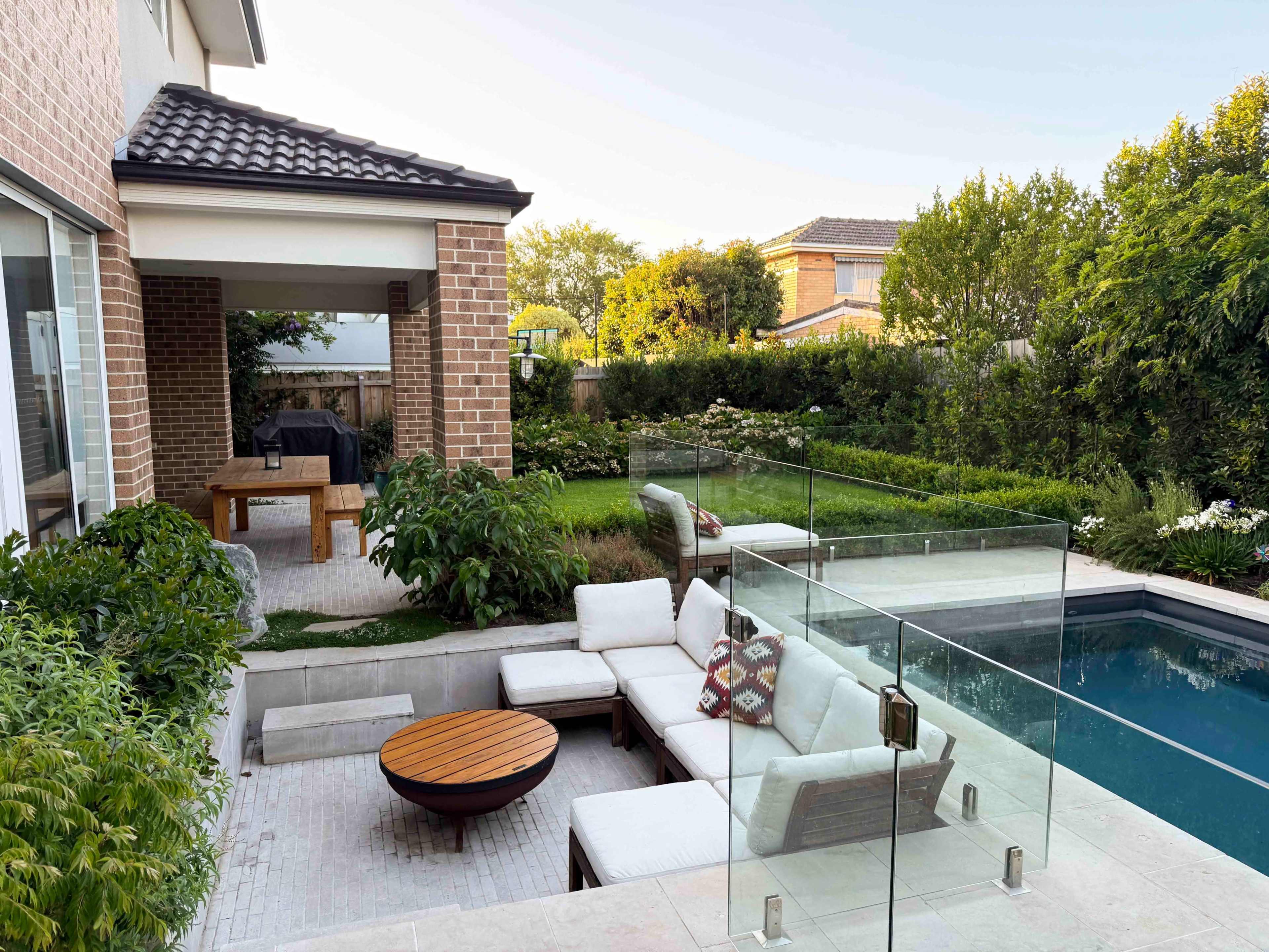 The image shows a modern backyard with a swimming pool, glass fencing, a sectional sofa, a circular wooden coffee table, and landscaped gardens.