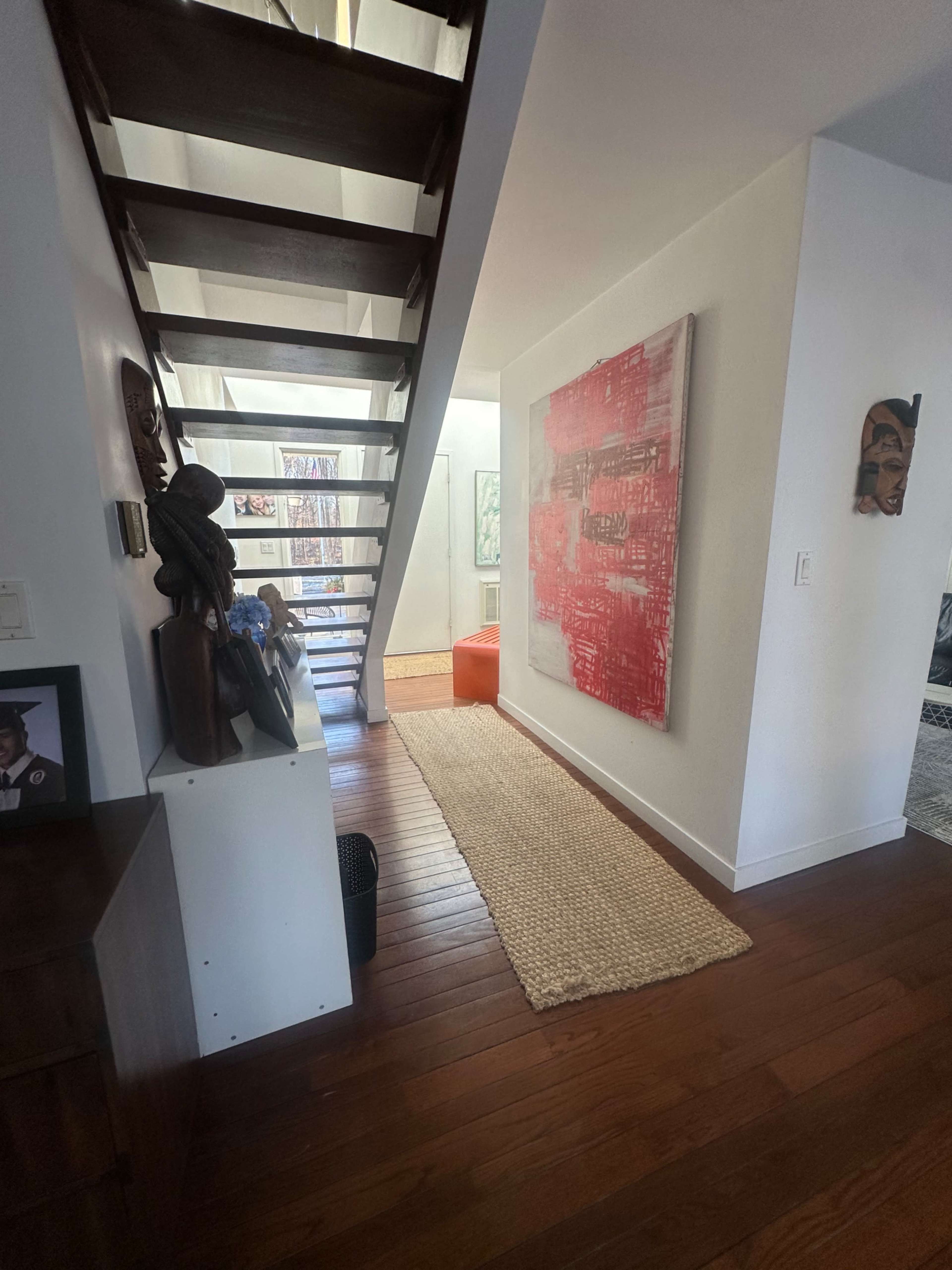 The image shows a modern hallway with a staircase on the left, colorful artwork on the wall, and a woven rug on the wooden floor.