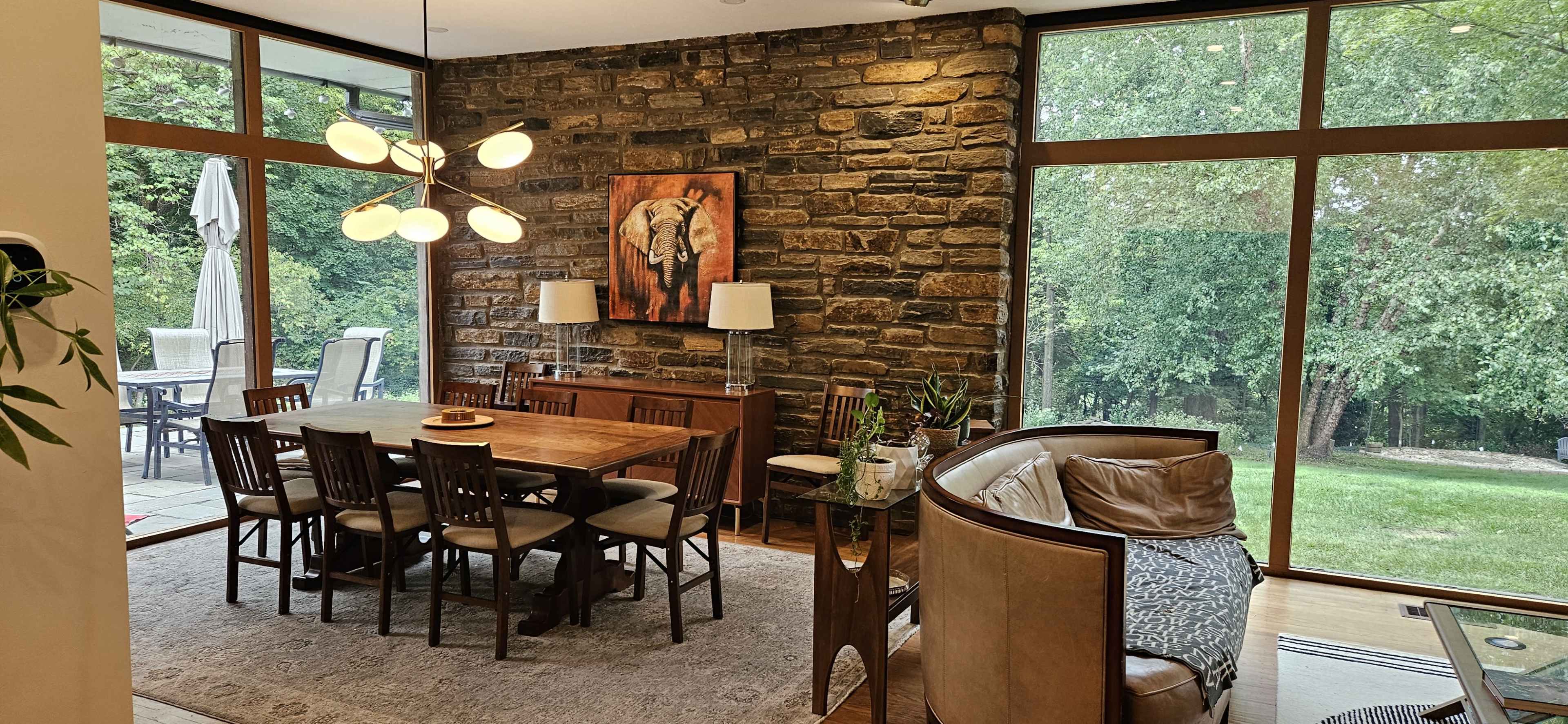A spacious dining area features a wooden table with chairs, surrounded by large windows and a stone wall, complemented by an elephant painting and modern light fixture.