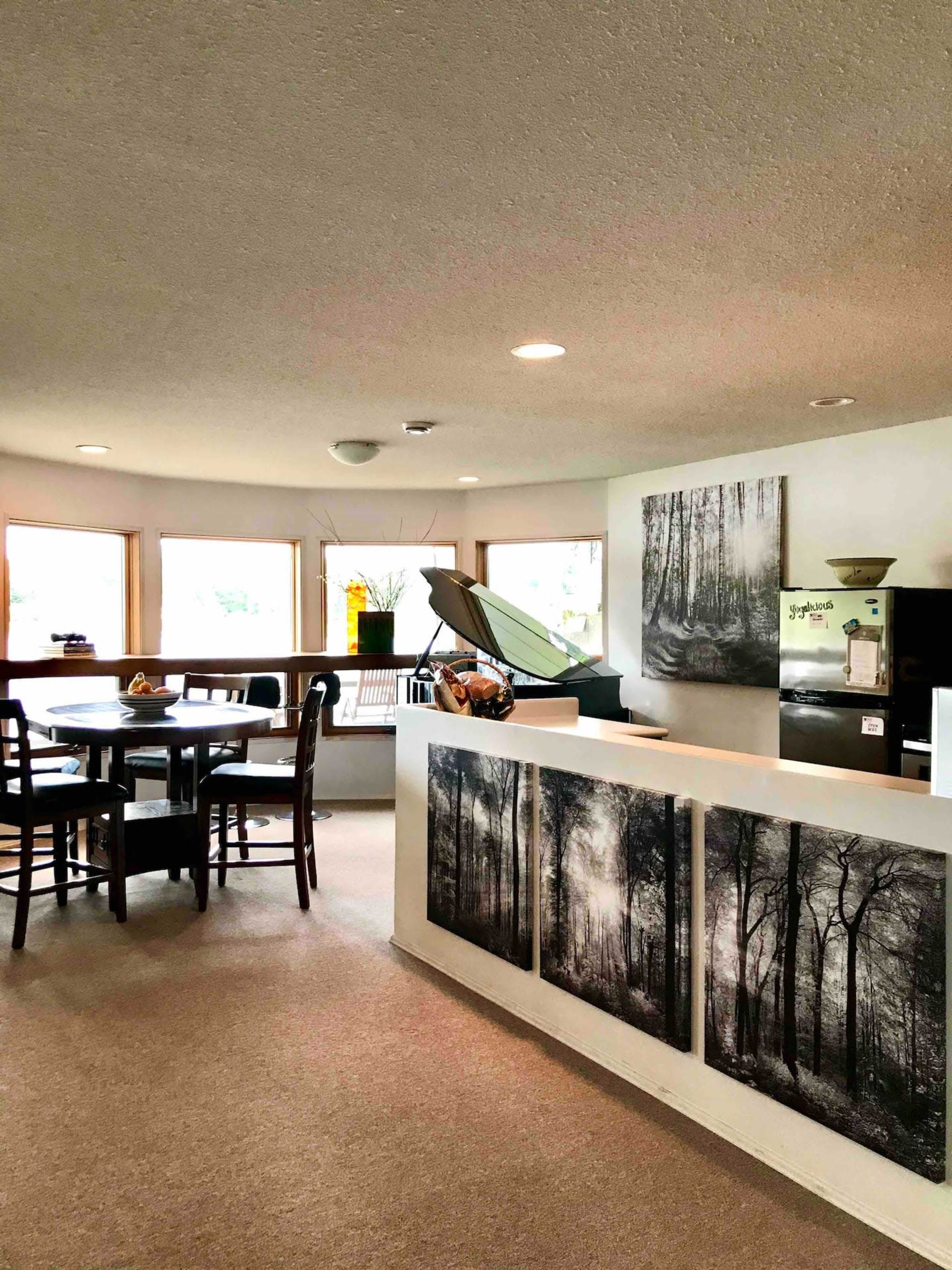 A well-lit, contemporary kitchen and dining area featuring a circular layout with a wooden dining table, chairs, and a wall decorated with black-and-white forest photographs.