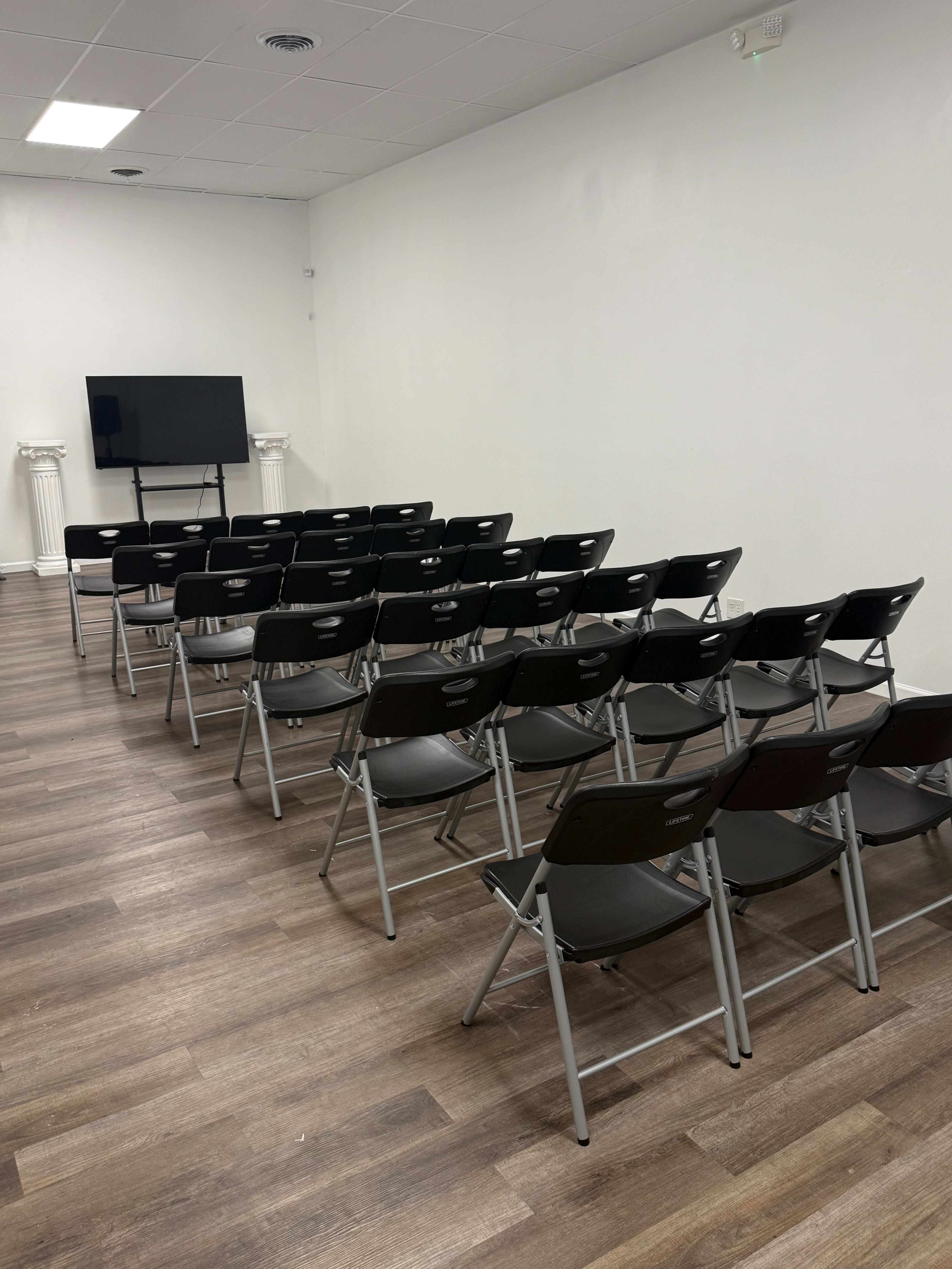 The image shows a meeting room with rows of black folding chairs facing a television mounted on a white wall.