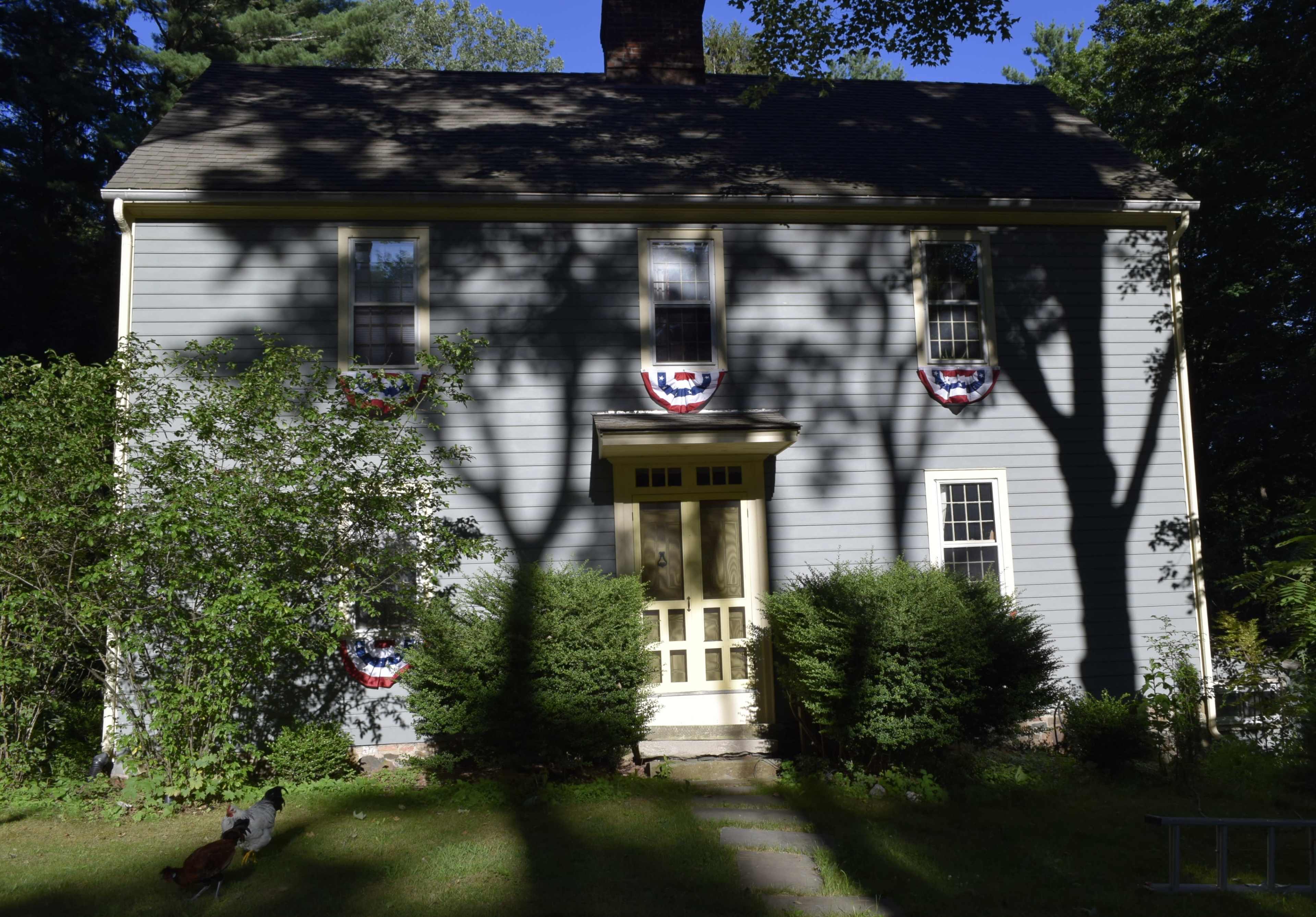A two-story gray house with patriotic bunting hanging on the front, surrounded by greenery and casting shadows on the lawn.