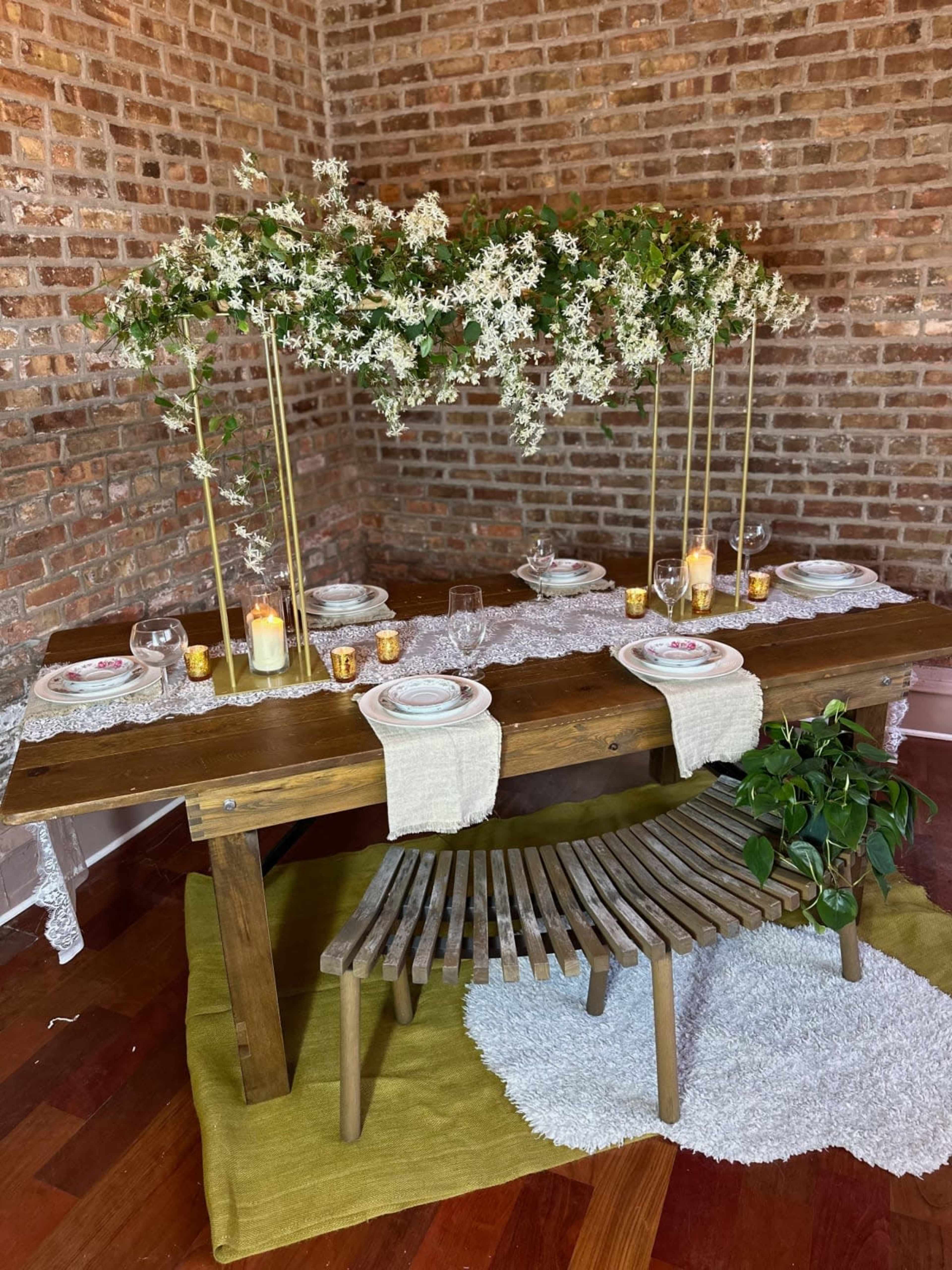 A wooden dining table is set with white plates, candles, and floral decorations above, surrounded by a brick wall and a green rug.