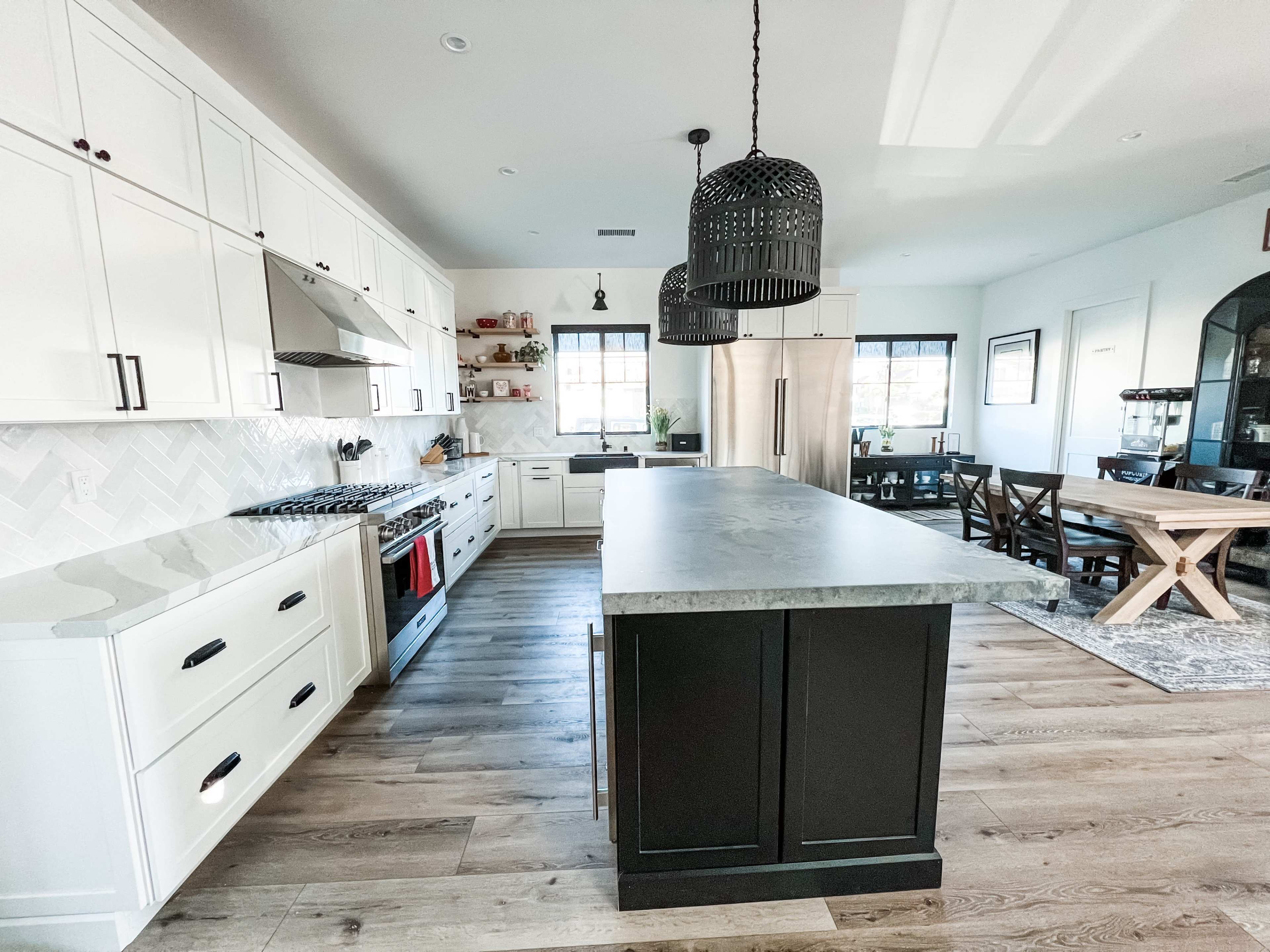 A modern kitchen features white cabinets, a large gray island, a gas range with a red accent, and a dining area with a wooden table.