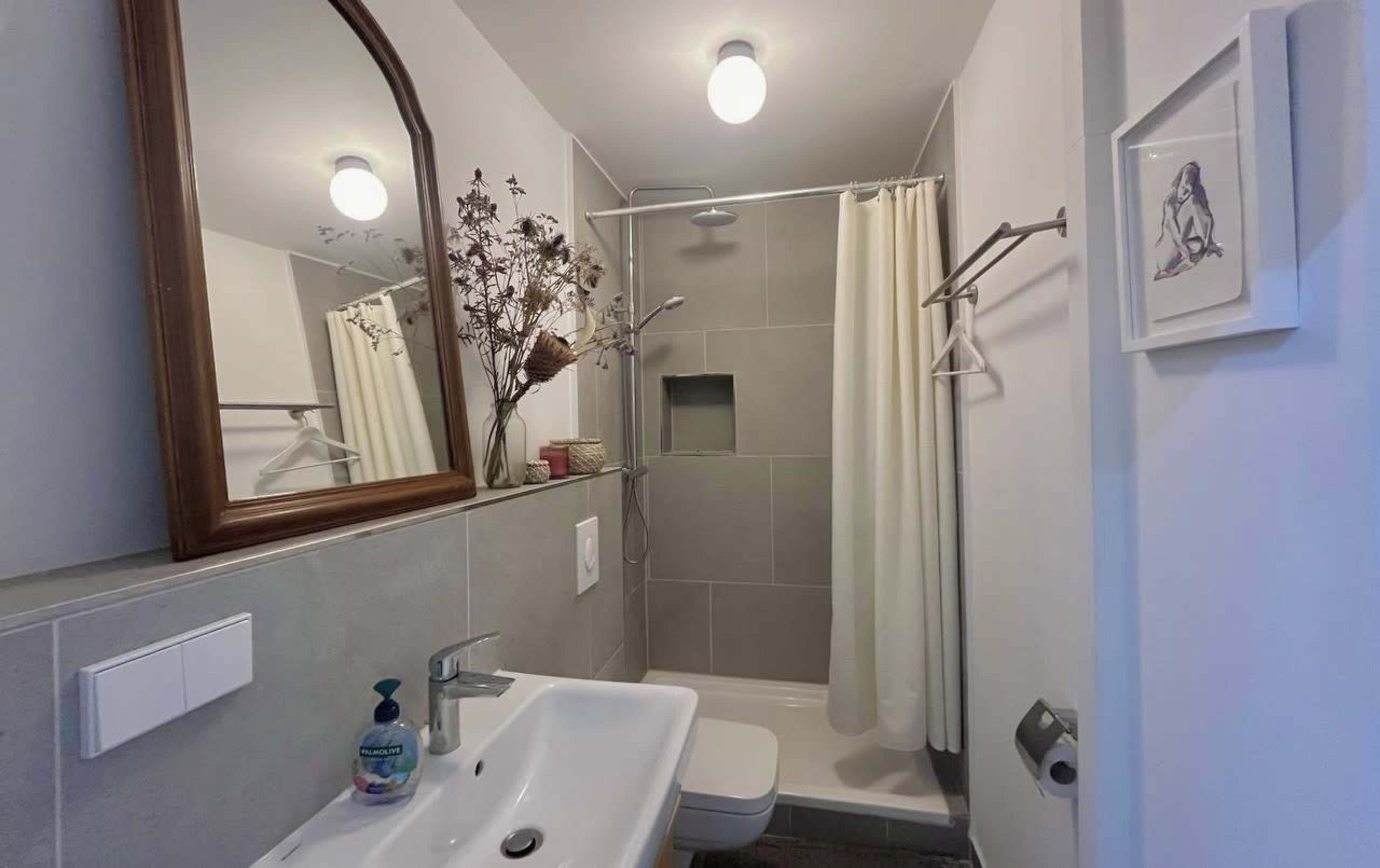 The image shows a modern bathroom featuring a shower with a curtain, a sink with a mirror above it, and a small decorative arrangement of dried flowers.