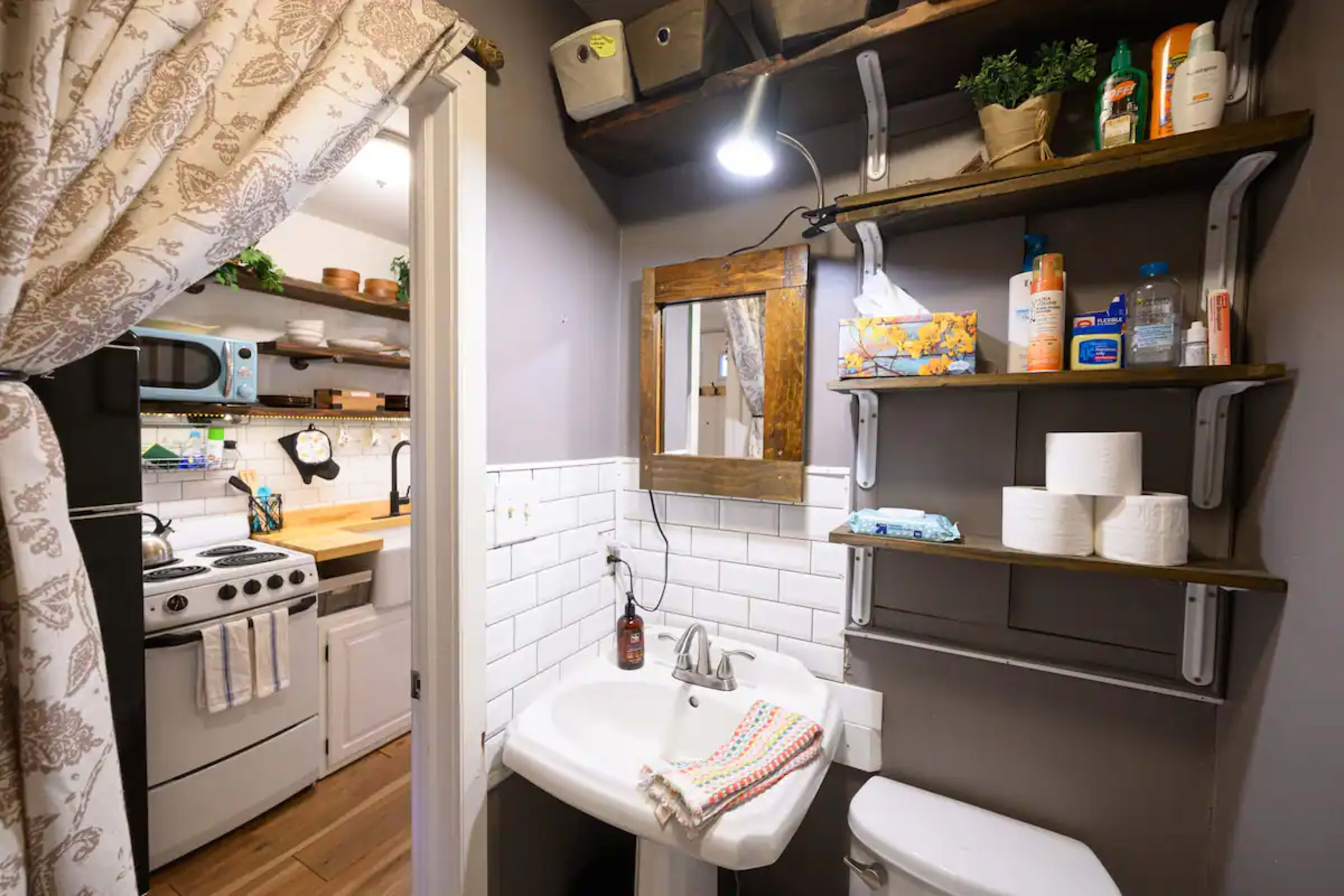 A small bathroom with a sink, toilet, shelves displaying toiletries, and a peek into a kitchen area with an oven.