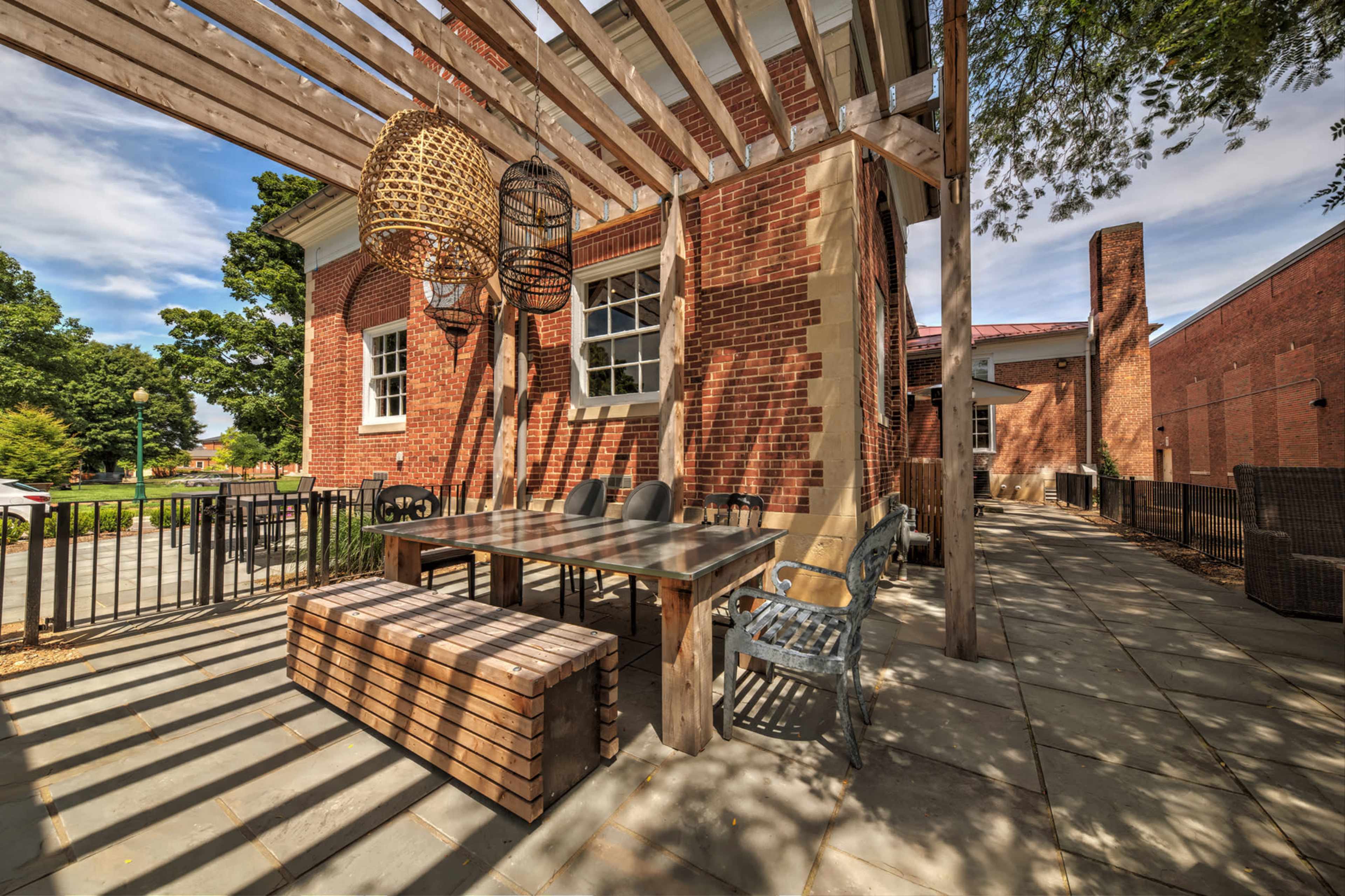 The image shows a brick patio area with a large wooden table and benches, shaded by a pergola and surrounded by greenery.