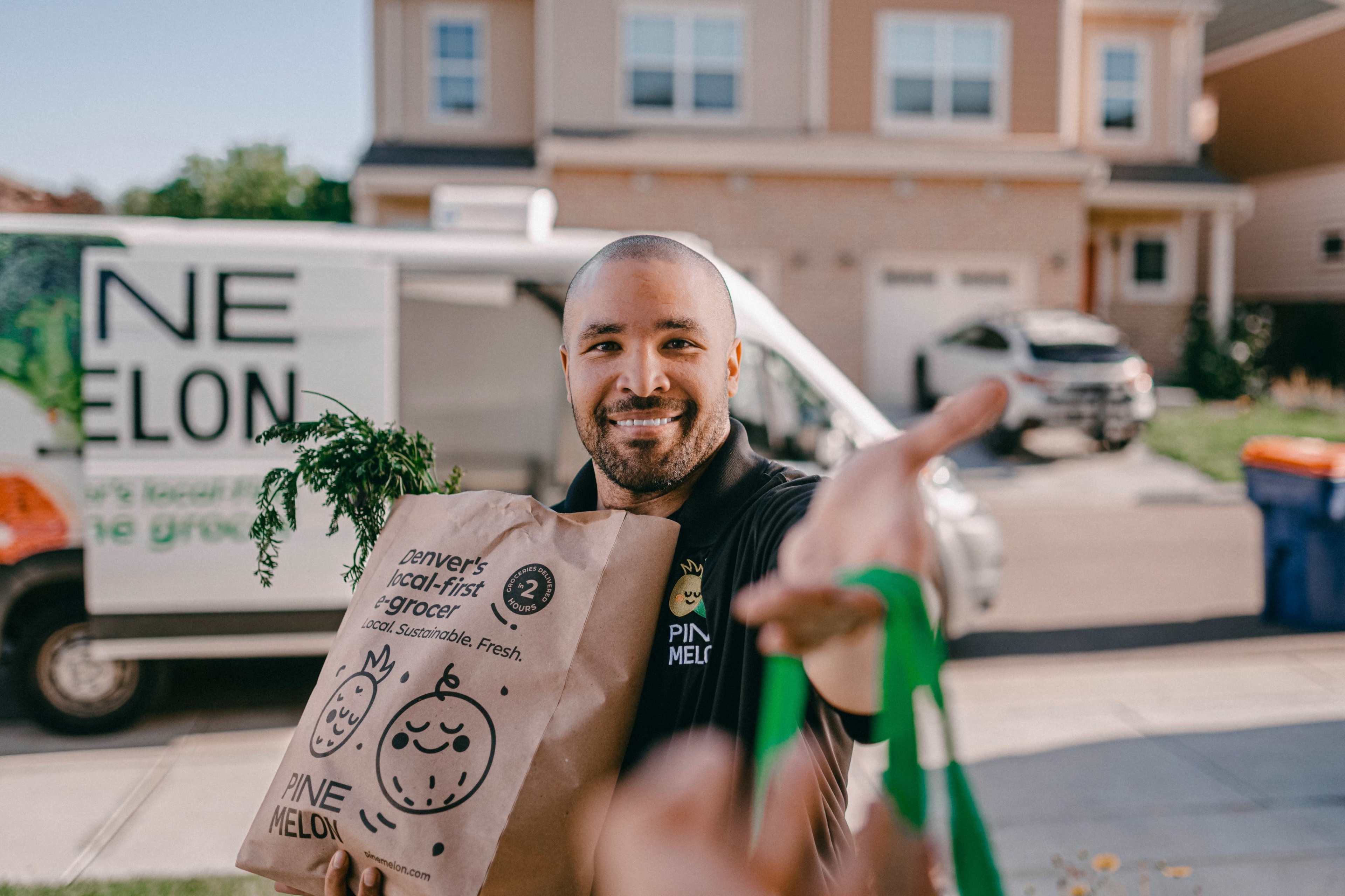 A man holds a brown grocery bag labeled "PINE MELON" while standing in front of a delivery van and a house.