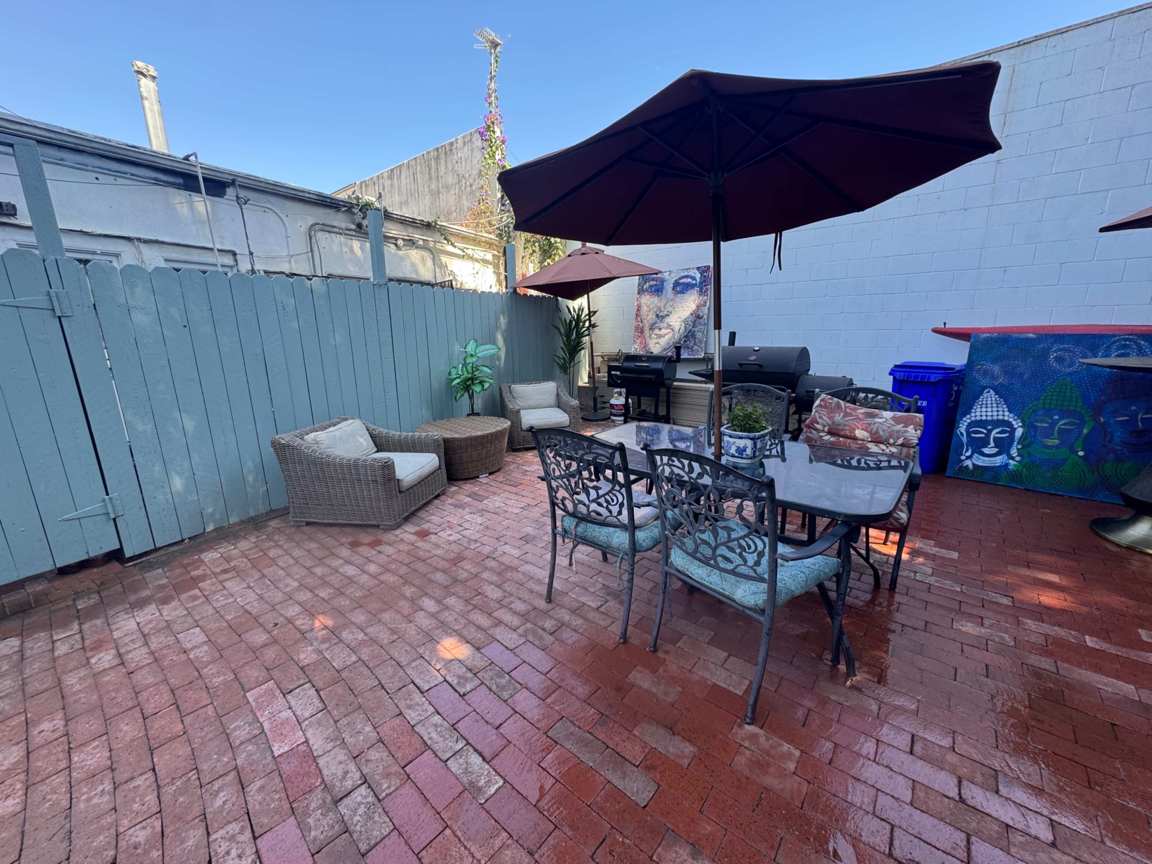 The image shows a brick patio area with outdoor furniture, including a dining table and chairs, a grill, and several potted plants, all shaded by an umbrella.