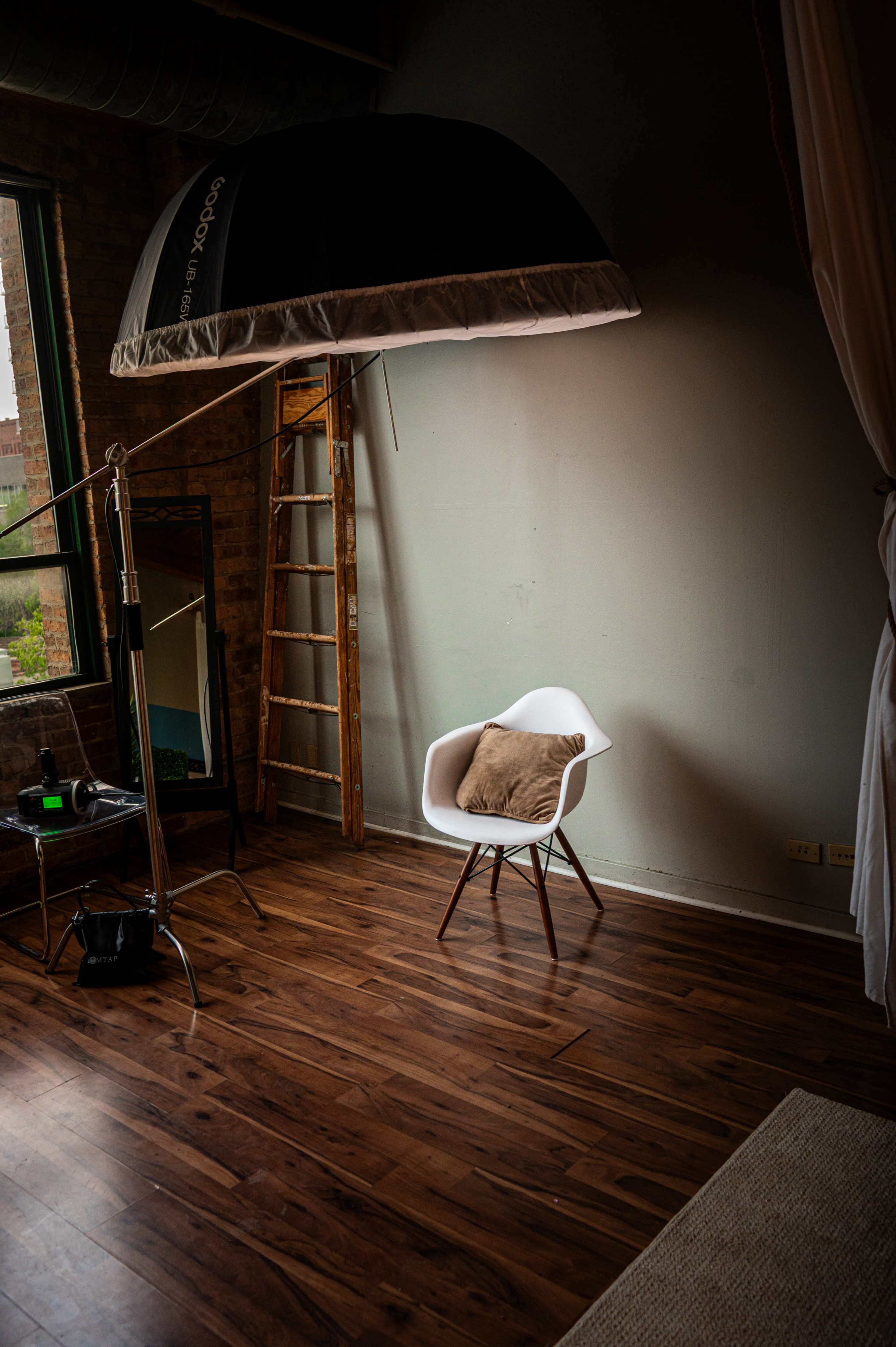 A large light umbrella is positioned above a minimalist white chair with a cushion in a well-lit photography studio.