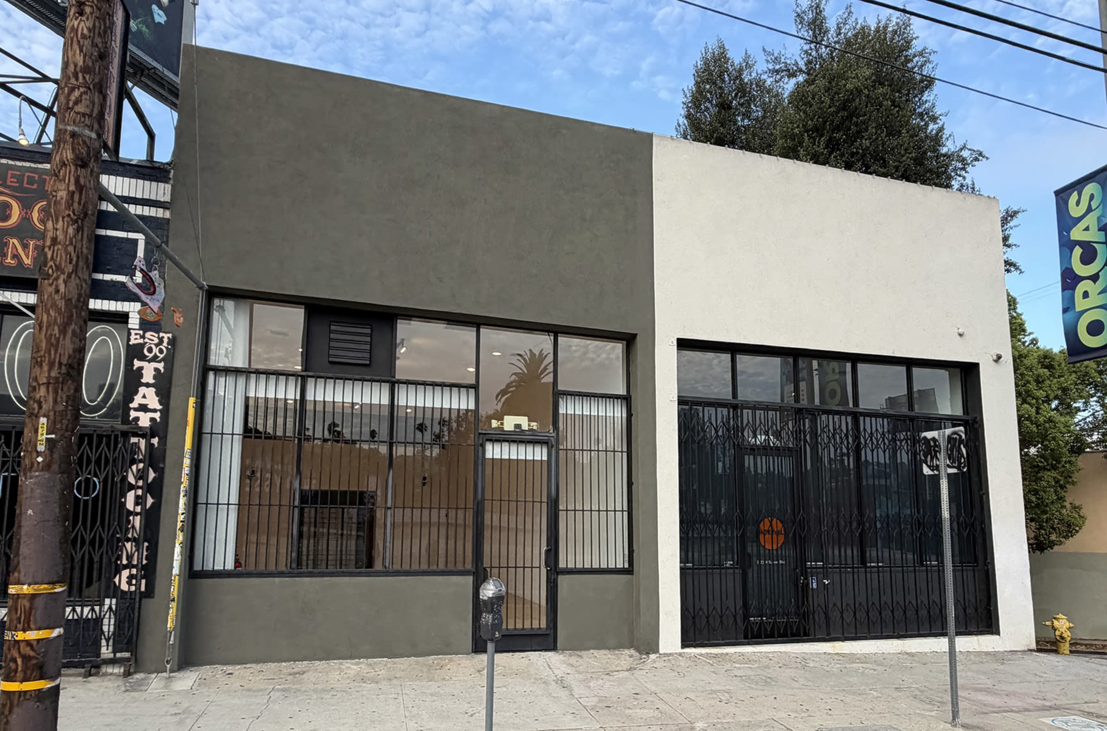 The image shows a commercial building with two distinct sections, one painted gray and the other white, featuring large windows and metal bars, situated on a sidewalk near utility poles and signs.