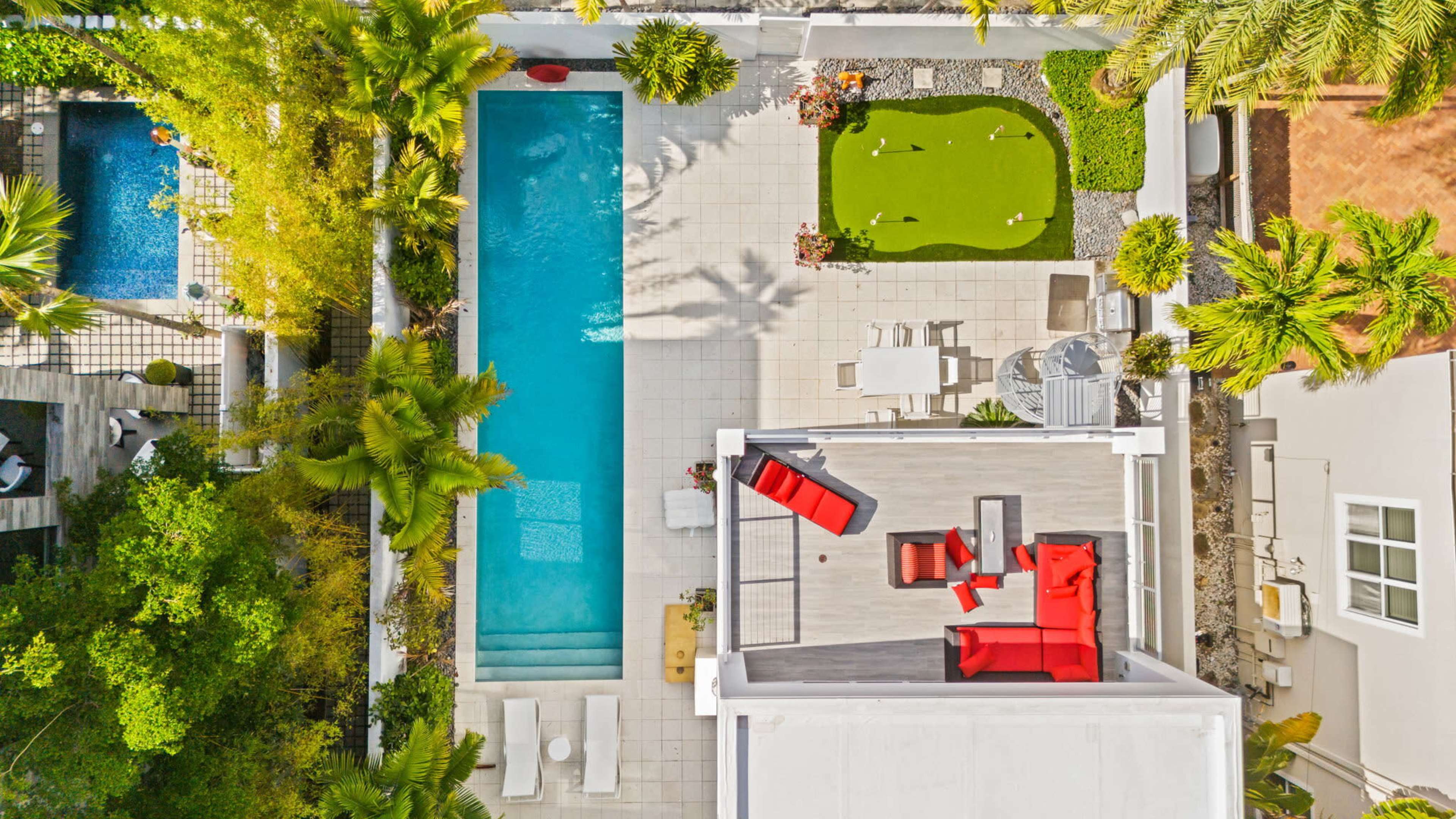 The image shows an aerial view of a modern backyard featuring a swimming pool, a manicured lawn, and outdoor seating areas surrounded by greenery.