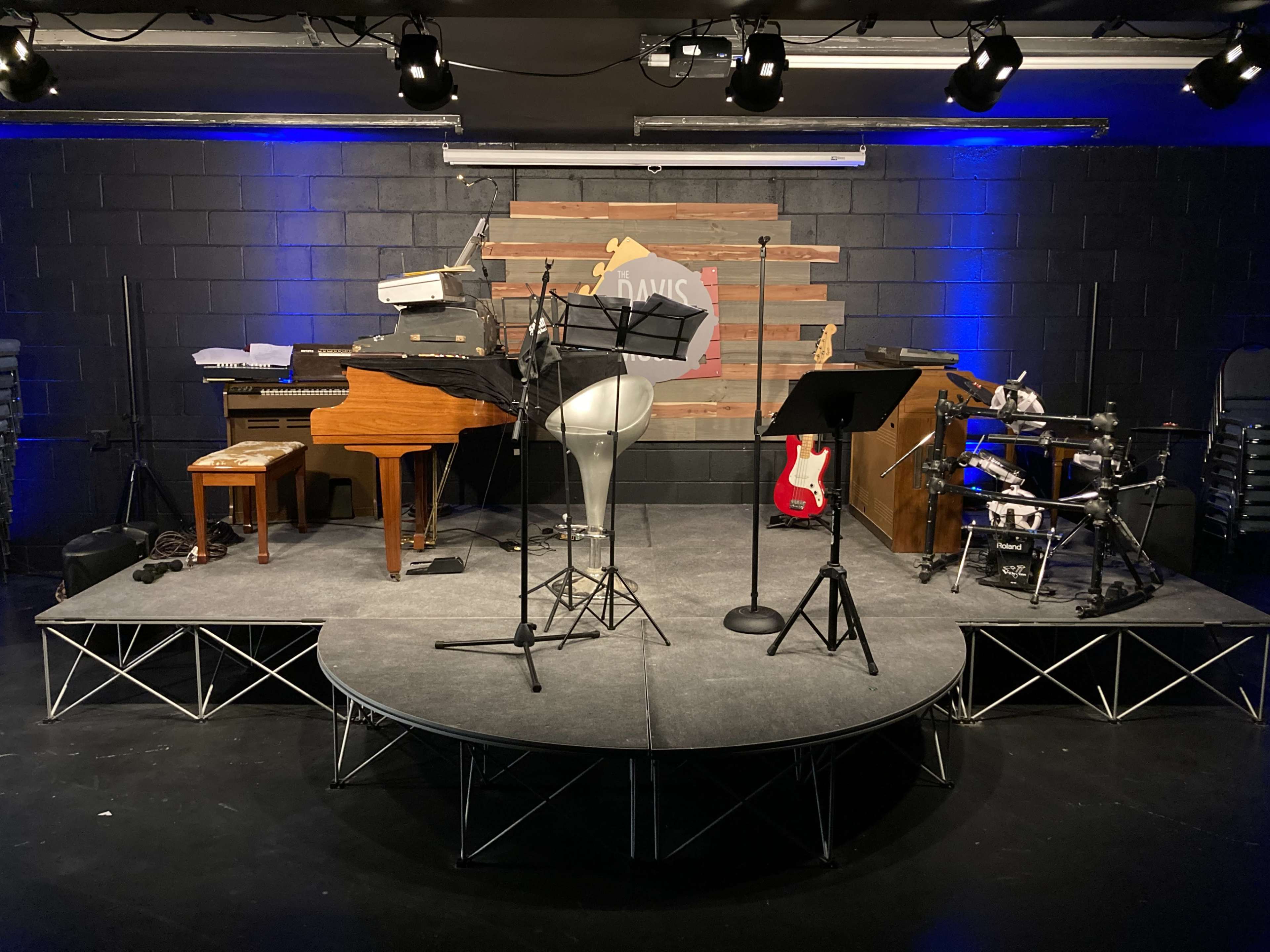 A small stage set up with a grand piano, a microphone stand, musical instruments, and music stands against a backdrop of wooden panels.