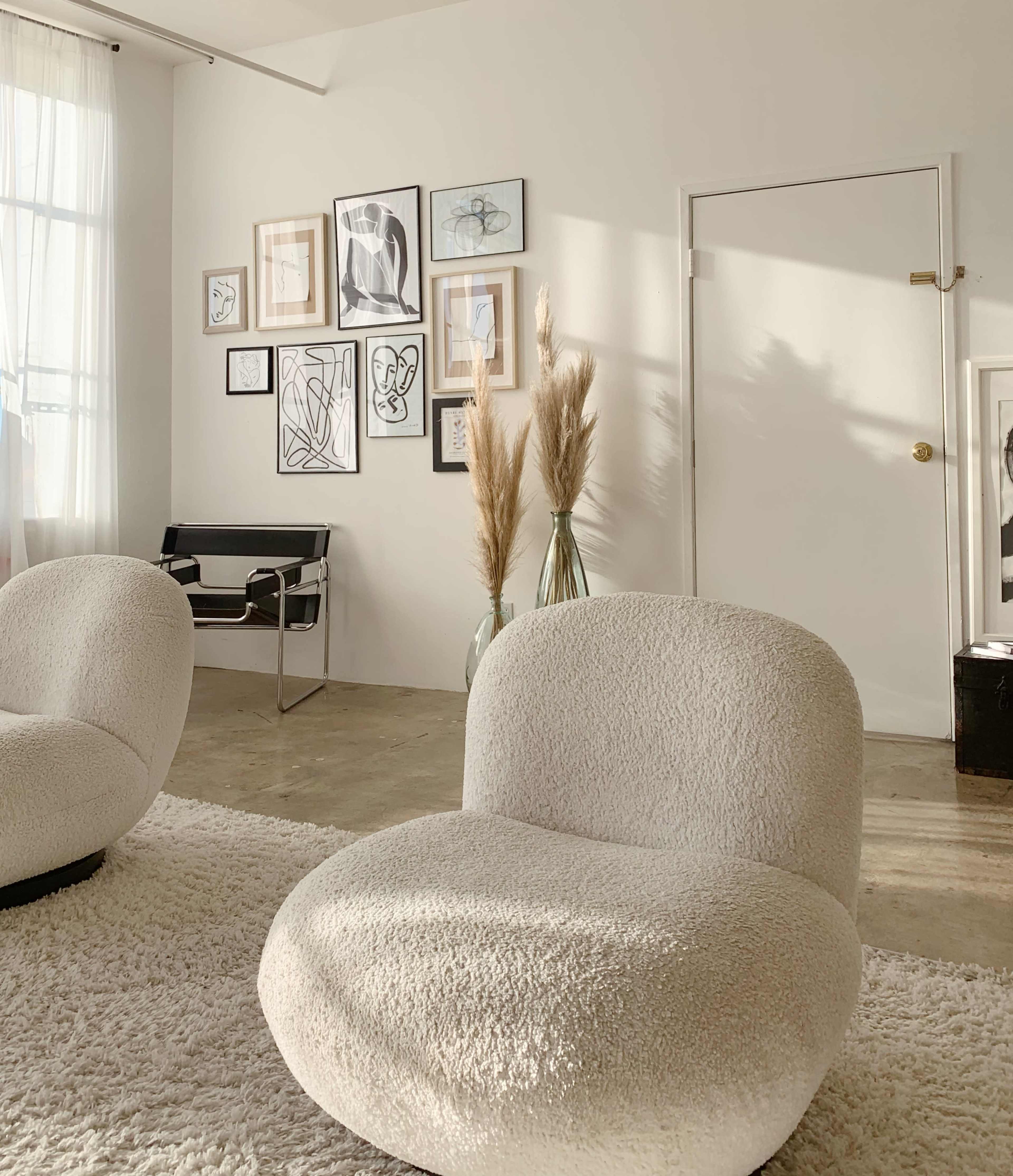 A minimalist interior with two white, rounded chairs facing a gallery wall of framed line art and a floor vase with dried pampas grass.