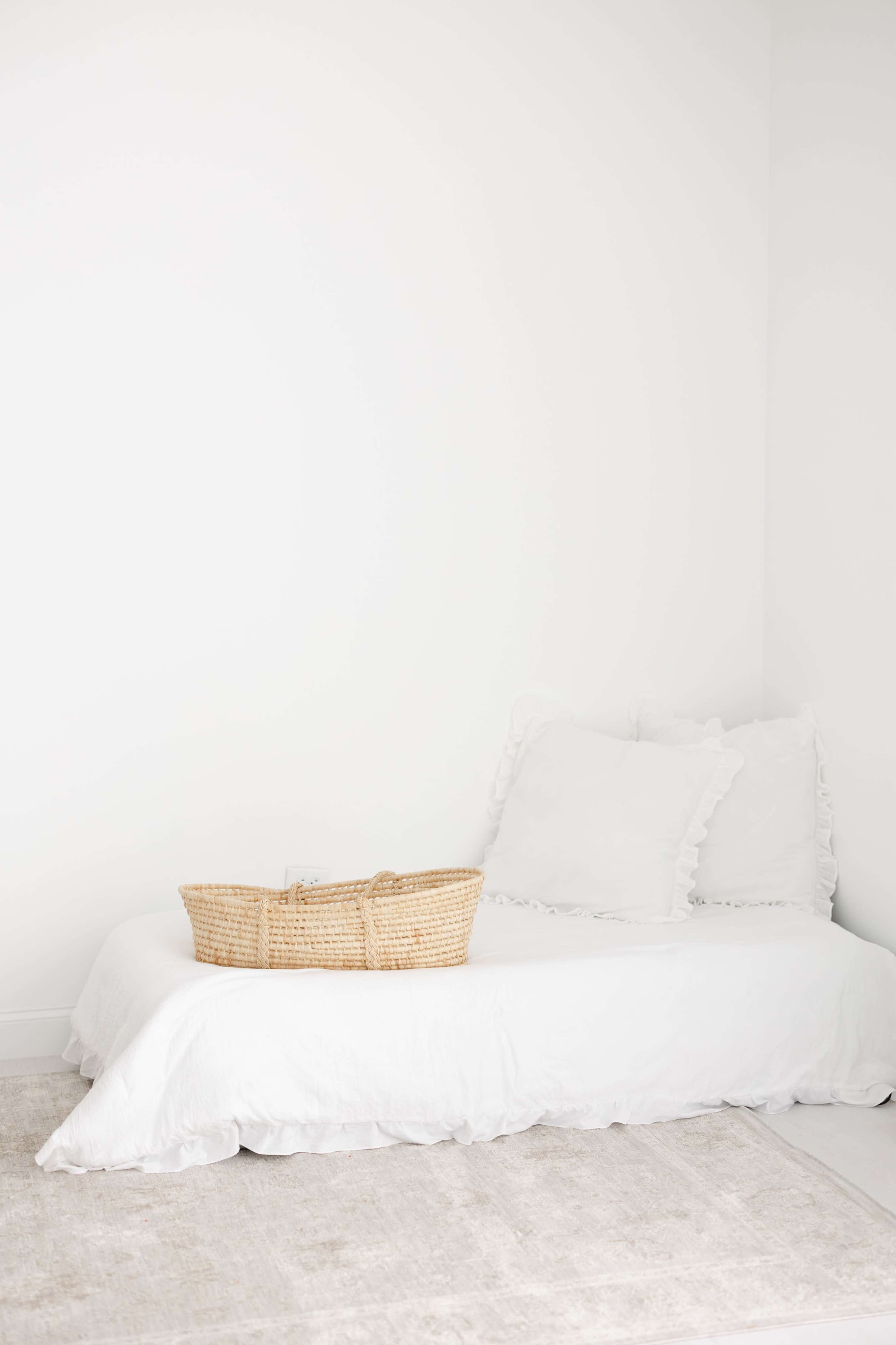 A simple bed with white bedding and two cushions is positioned against a plain white wall, accompanied by a woven basket.