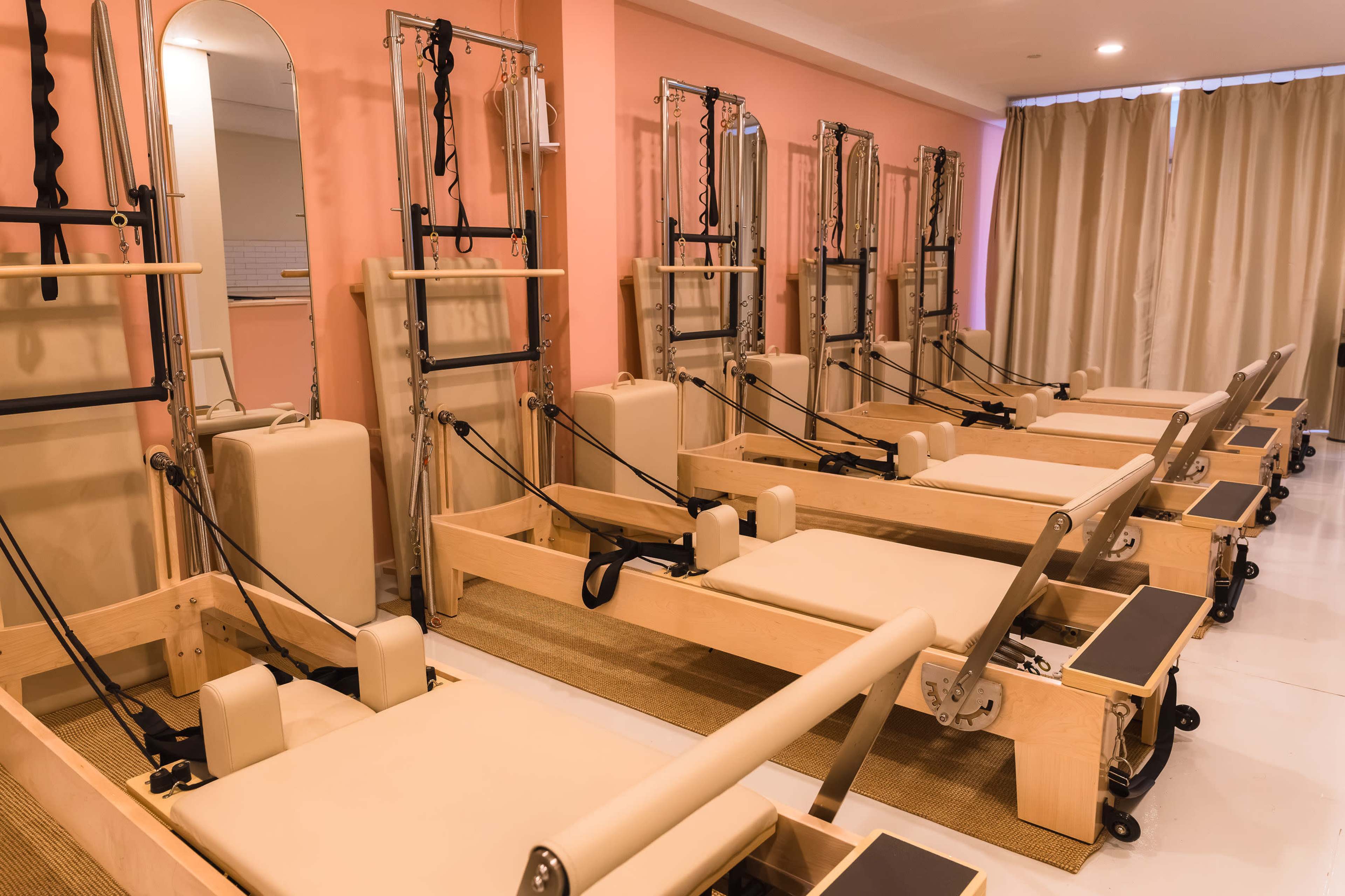 The image shows a series of reformer Pilates machines arranged neatly in a bright studio with pale pink walls and beige curtains.