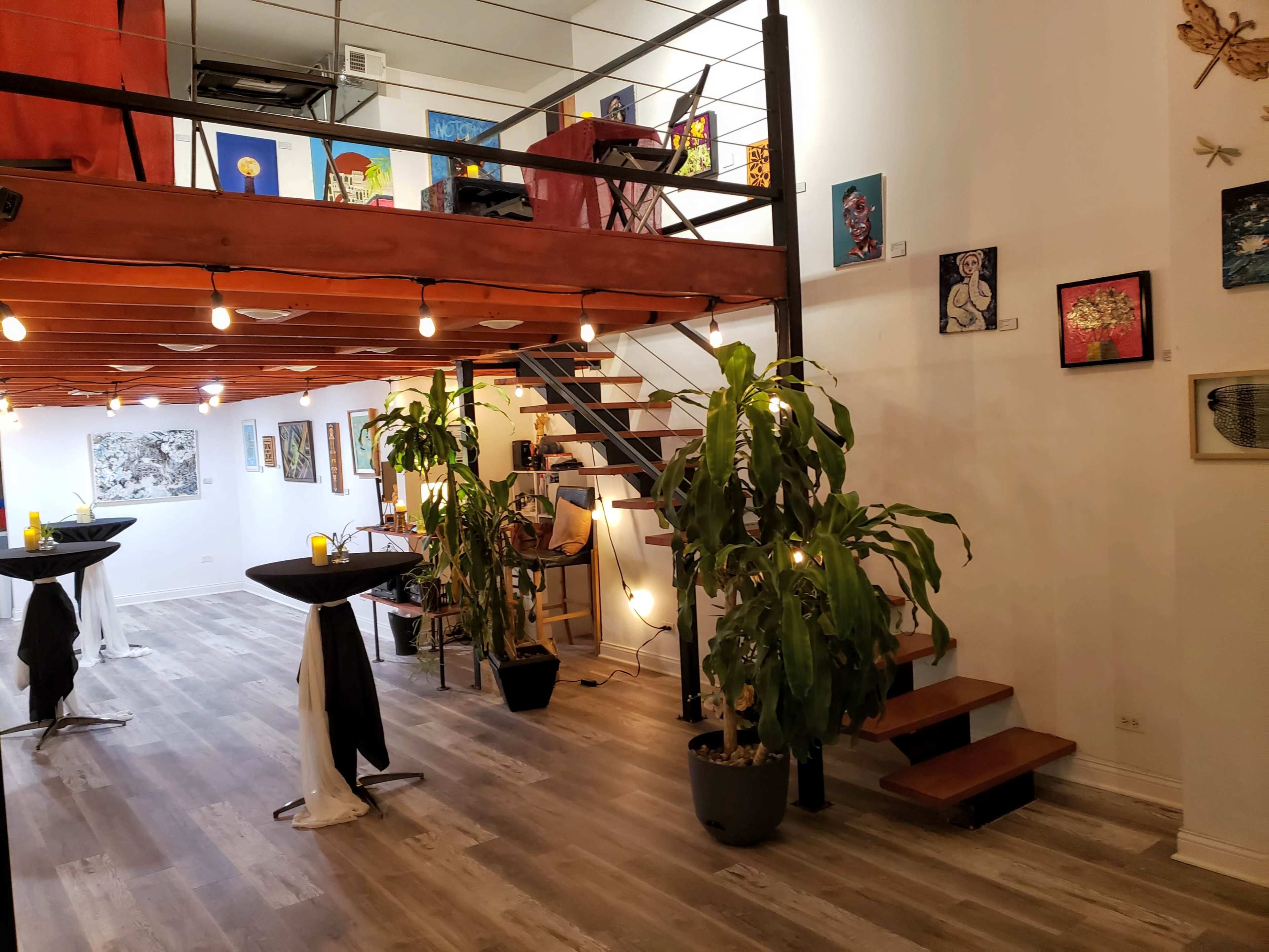 The image shows a spacious, art-decorated room with a staircase leading to a loft area, featuring plants and tables arranged for an event.