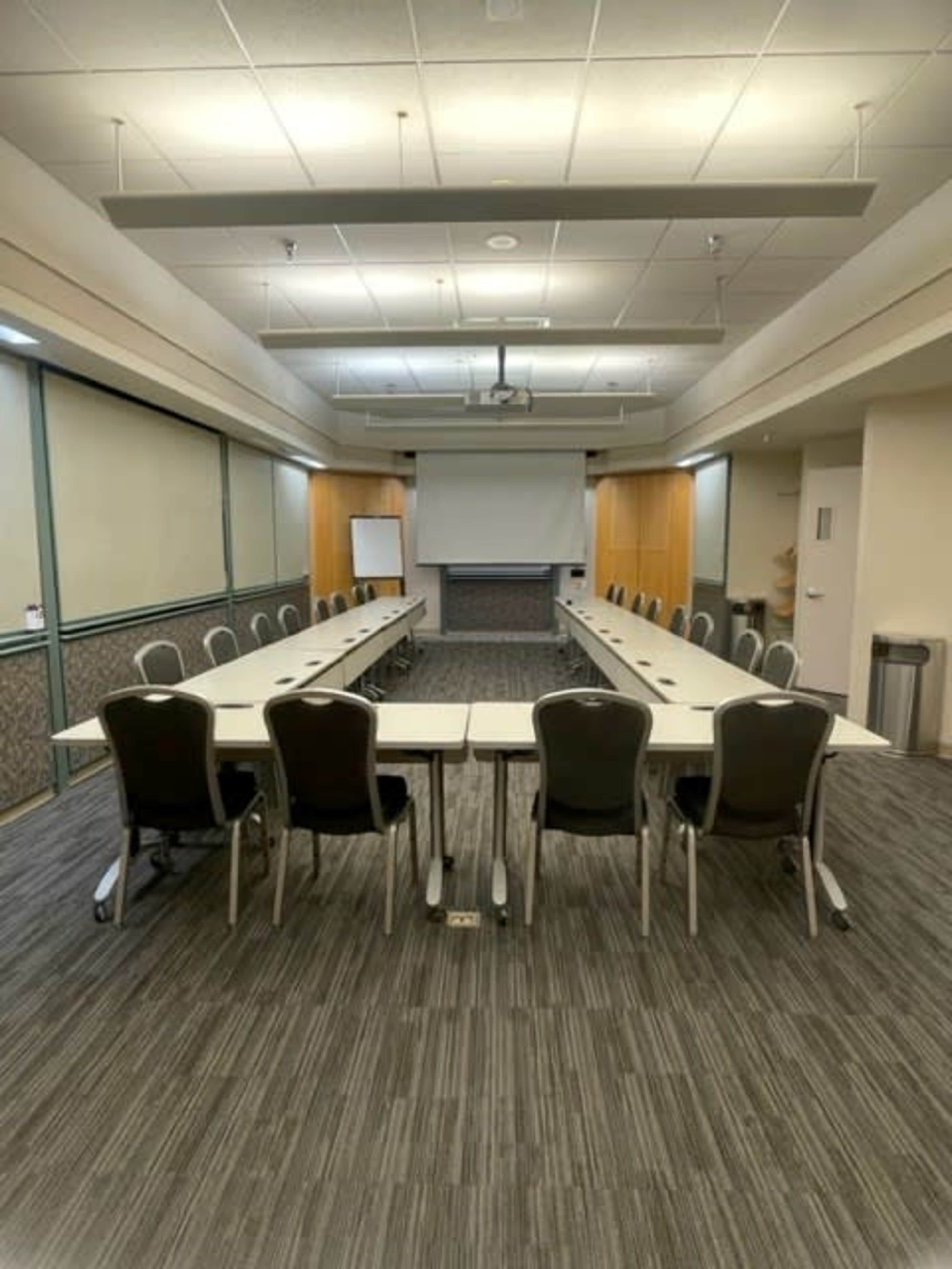A conference room is set up with a long rectangular table surrounded by chairs, with a projector screen and whiteboard at one end.