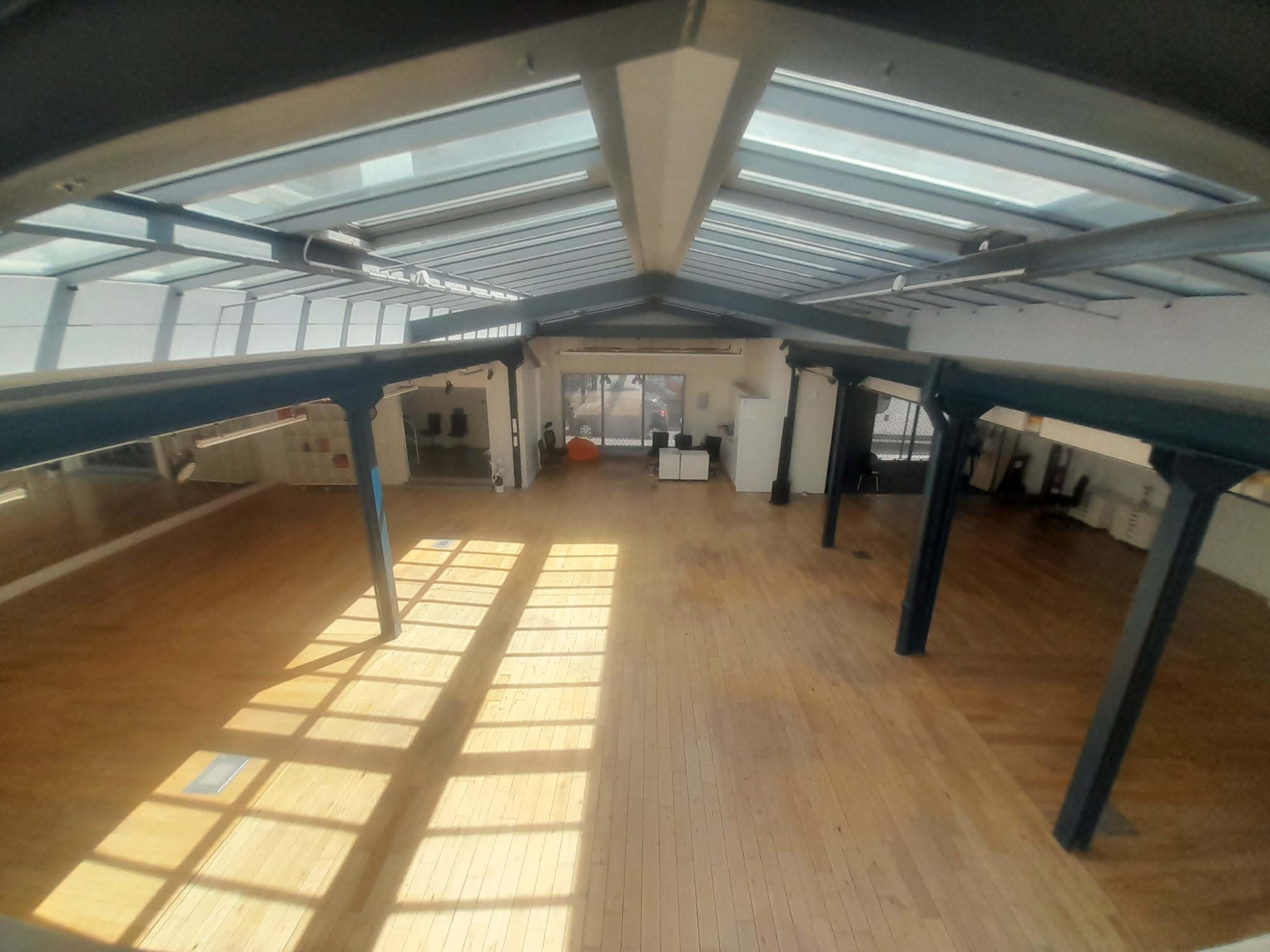 The image shows a spacious, well-lit interior with wooden flooring and large windows, featuring pillars and a minimalist design.