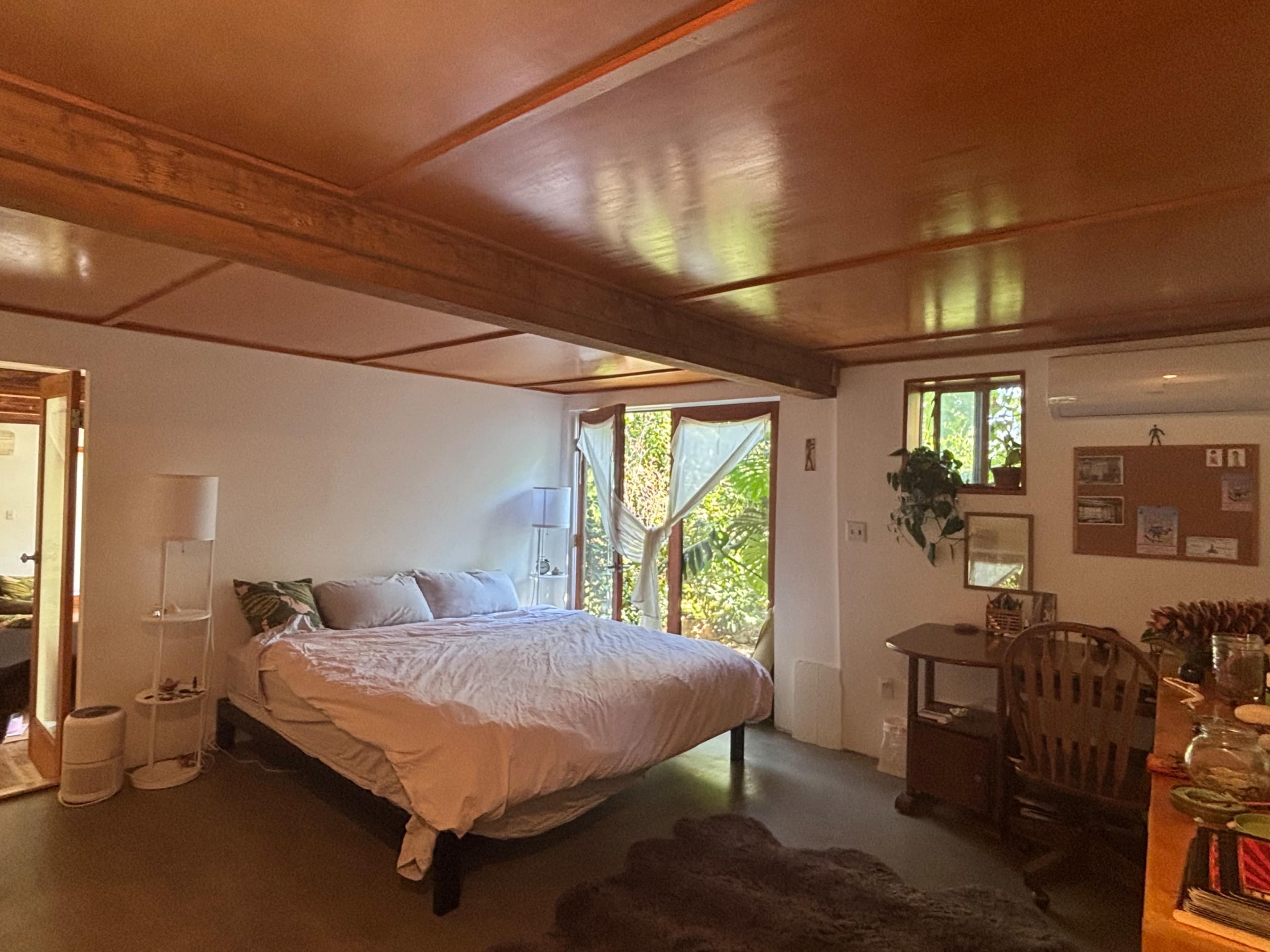 The room features a large bed, a writing desk, and a window with natural light illuminating the space, surrounded by greenery.