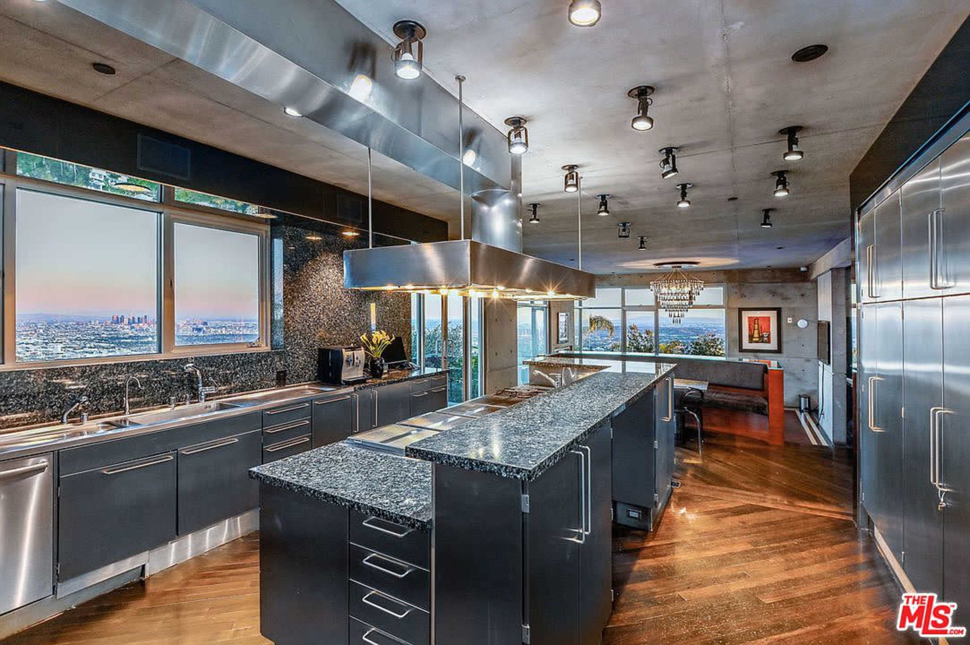 The image shows a modern kitchen featuring stainless steel appliances, dark cabinetry, and a central island with a view of the city through large windows.