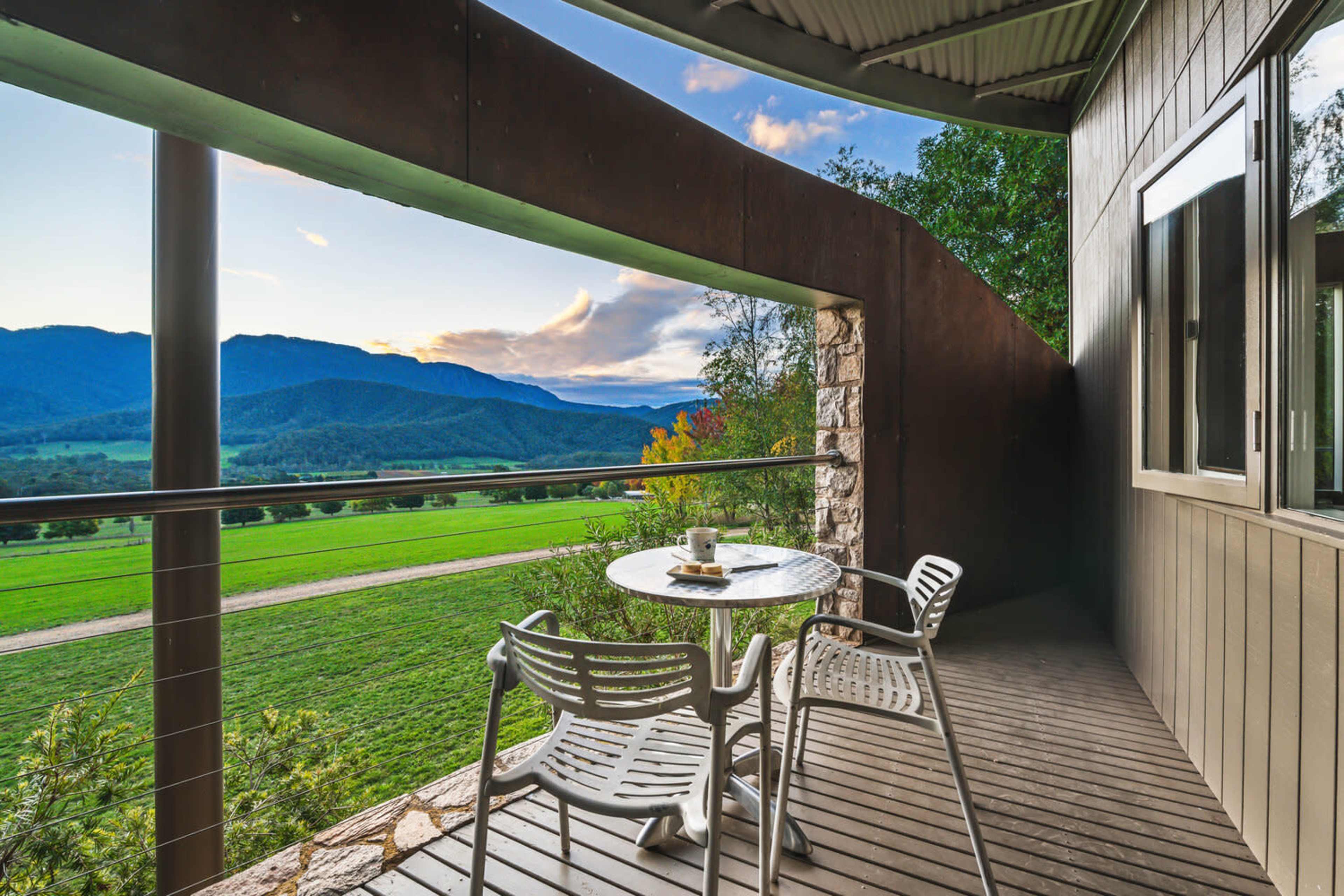 A small balcony features a table and two chairs overlooking a green valley and distant mountains as the sun sets.