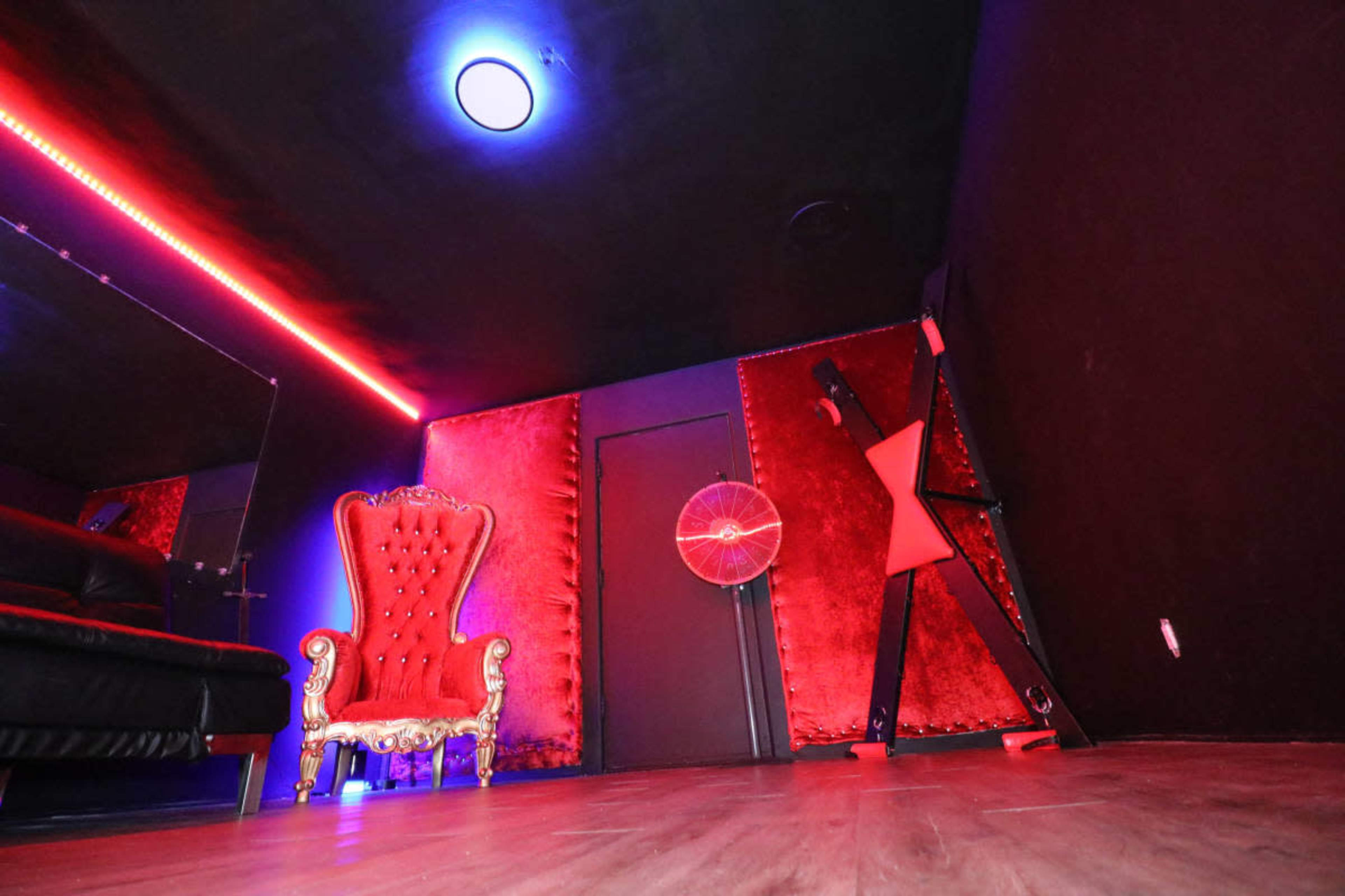 The image shows a dimly lit room featuring a large red velvet armchair, a black leather sofa, and illuminated red and blue neon lights along the walls.