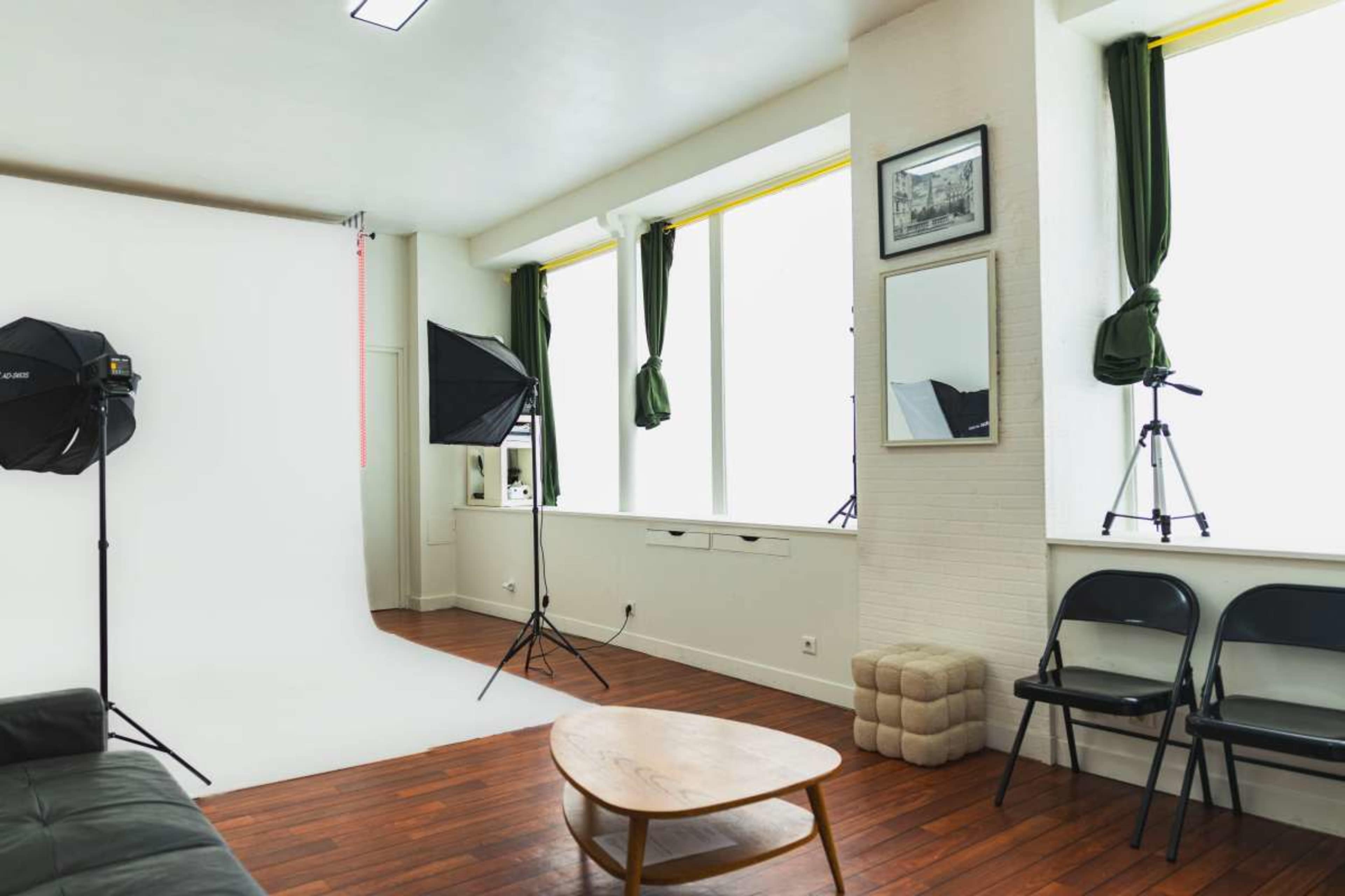 The image shows a photography studio featuring a white backdrop, softbox lights, a mirror, and a set of black folding chairs.