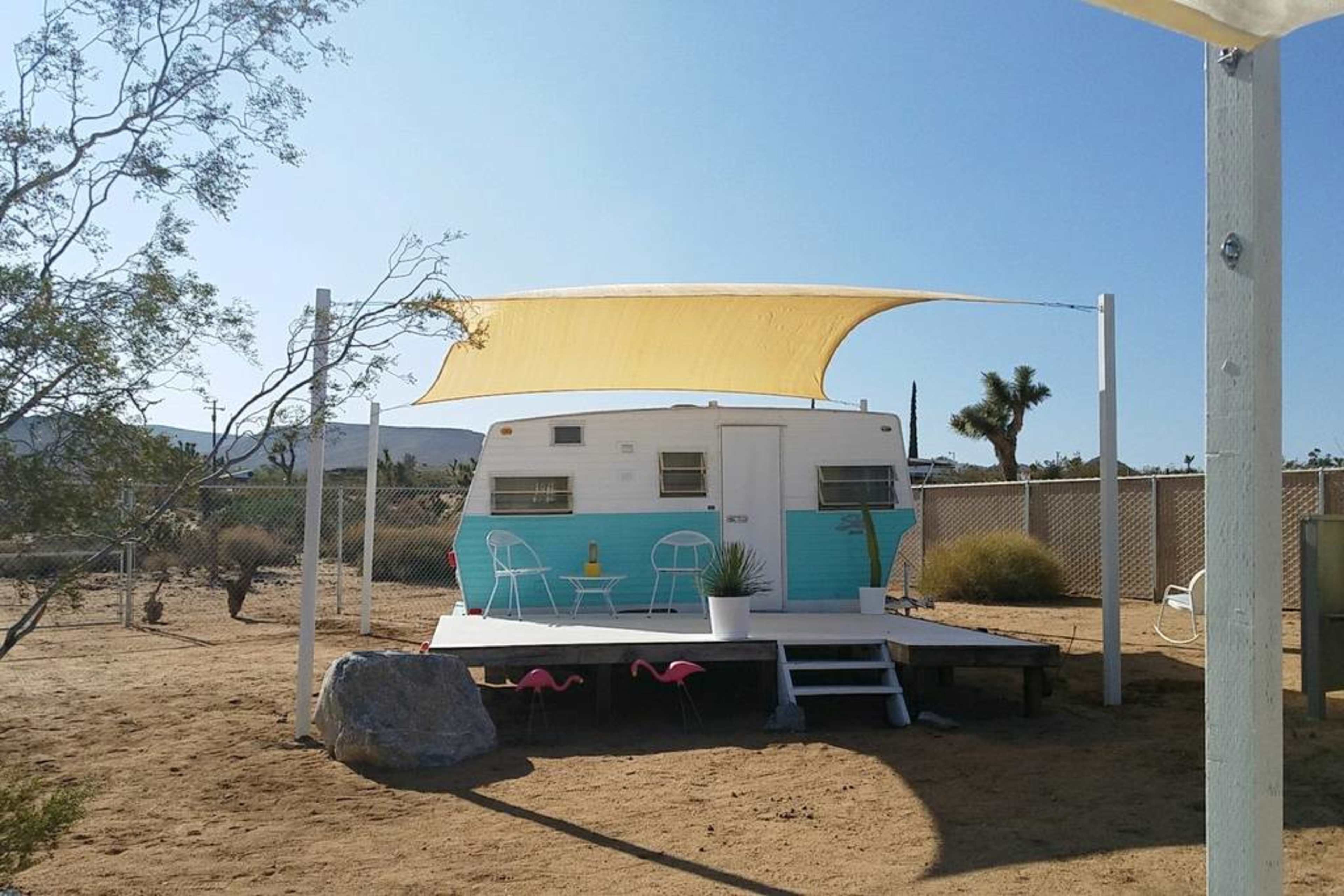 A vintage trailer with turquoise accents is positioned on a wooden platform, shaded by a yellow canopy, surrounded by desert landscape and decorative flamingos.