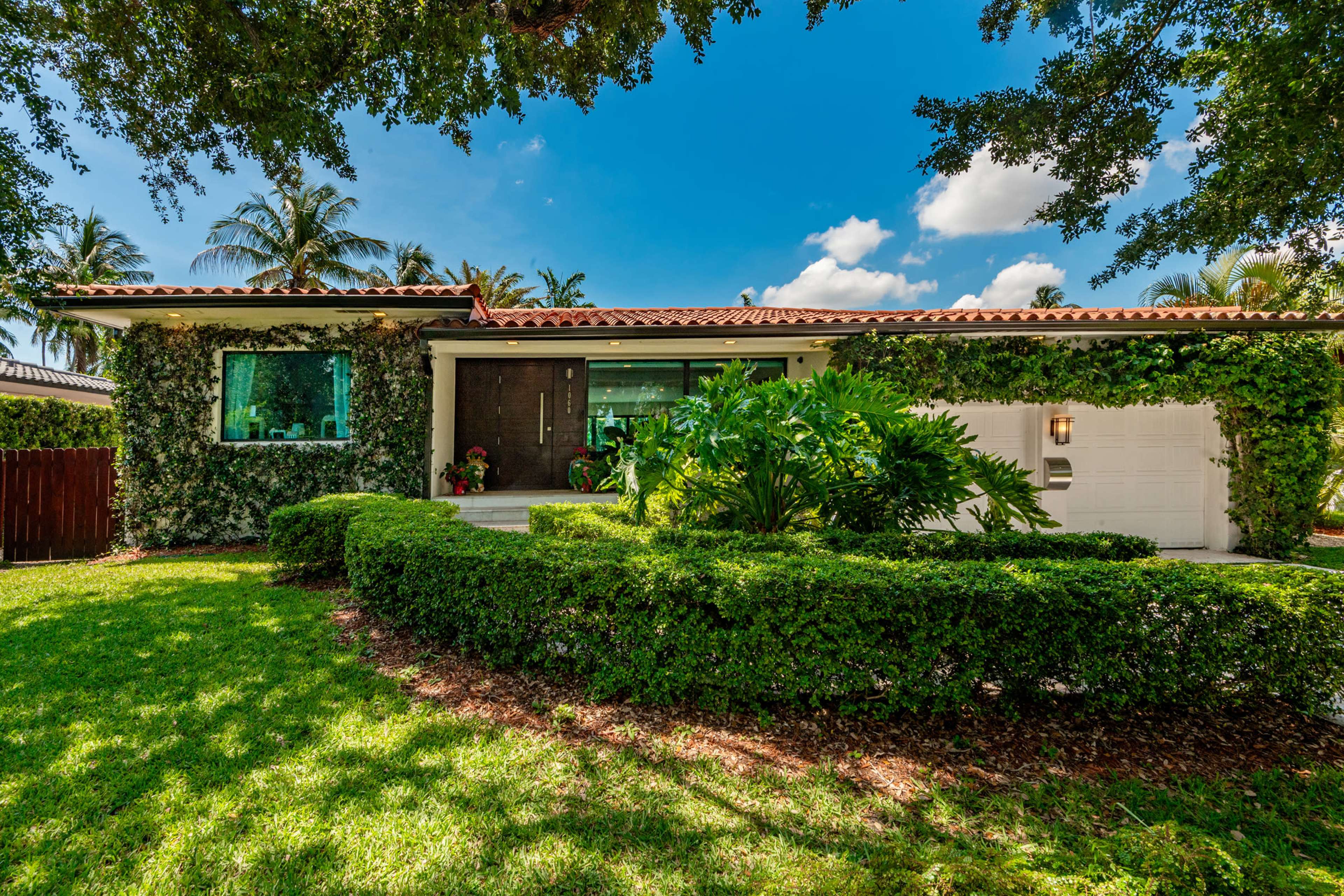 A modern house with a landscaped front yard featuring green shrubs and palm trees under a blue sky.