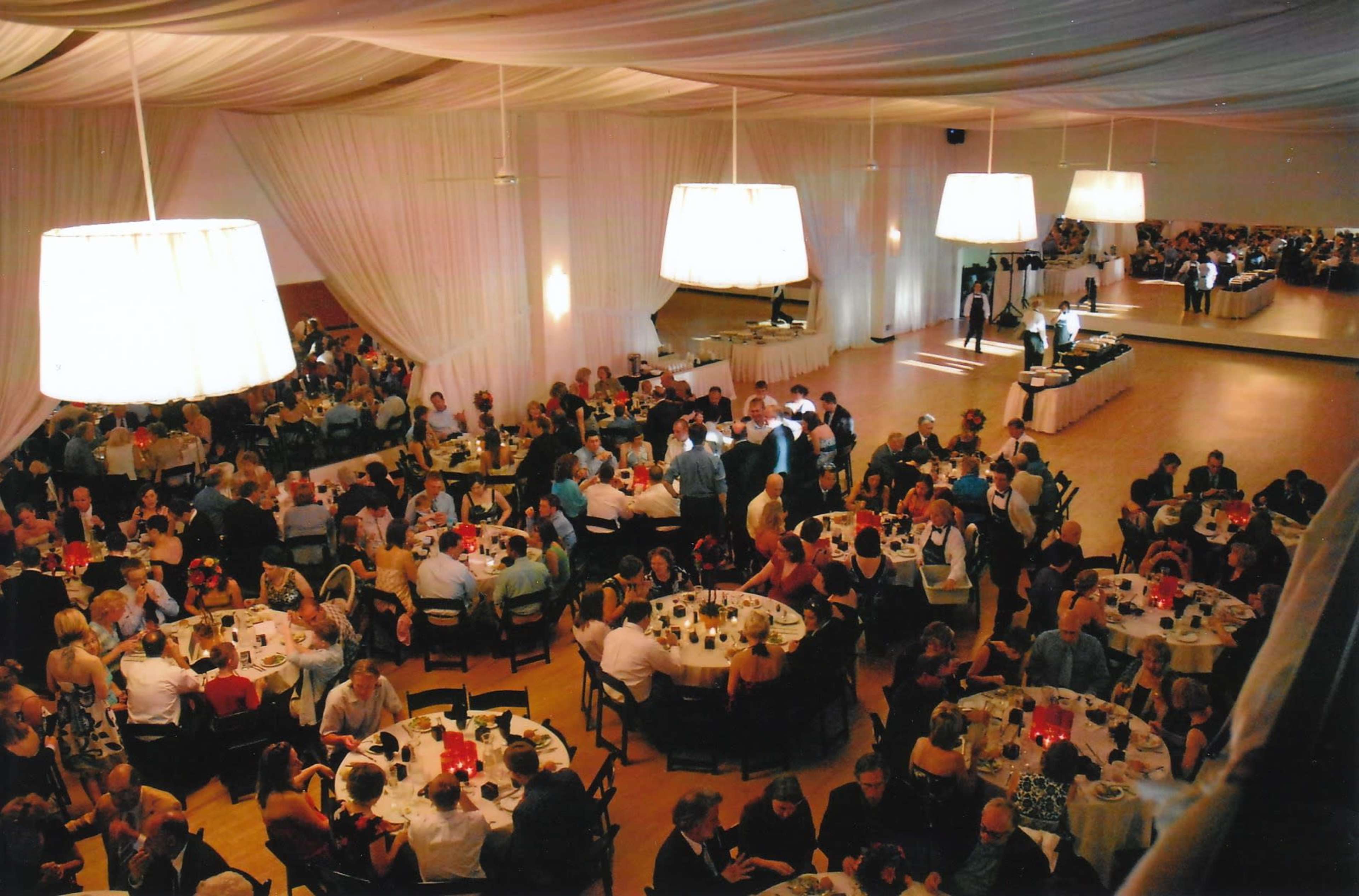 A large banquet hall is filled with numerous guests seated at round tables adorned with white tablecloths and lit candles, while a band plays in the background.