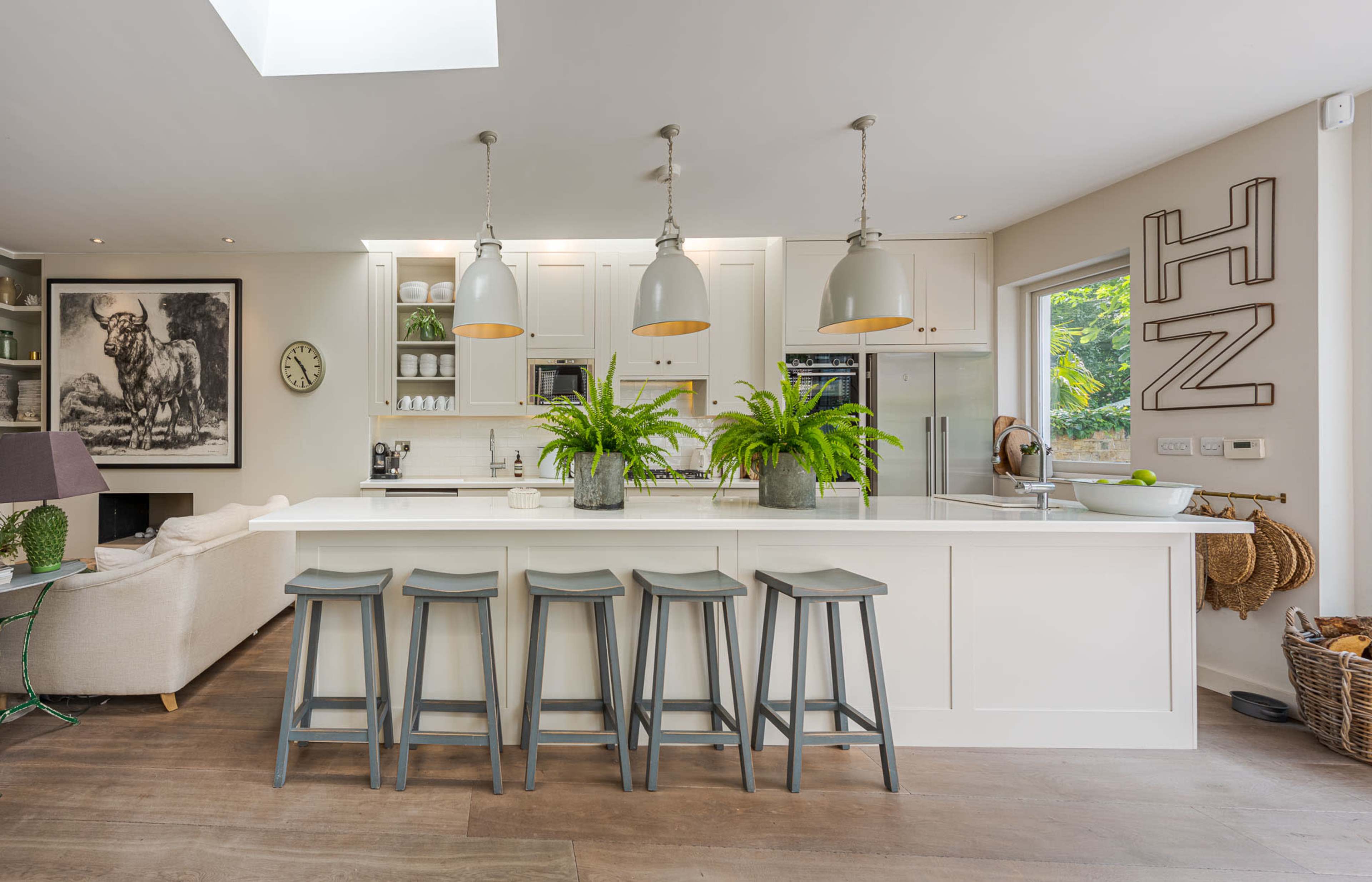 A modern kitchen features a central island with four gray stools, pendant lights overhead, and lush green plants on the countertop.