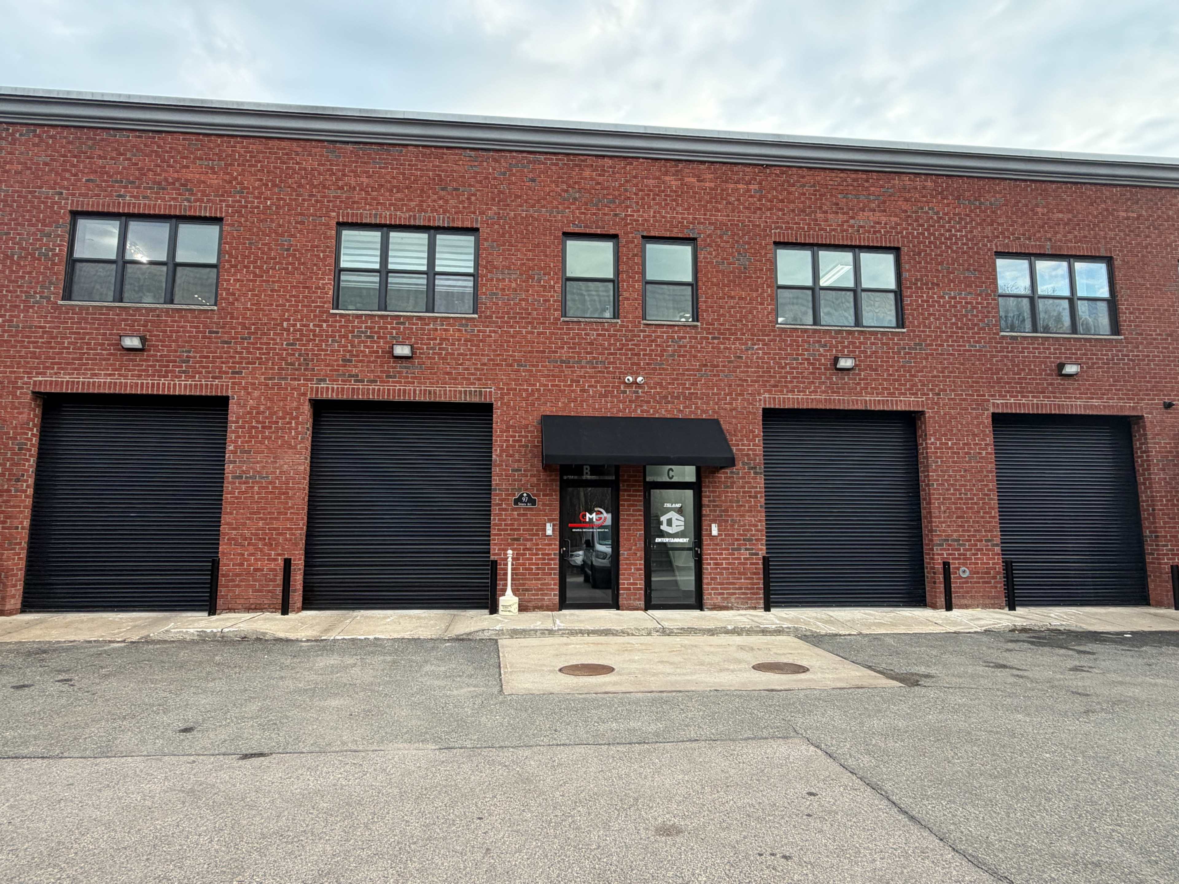 The image shows a red brick commercial building with three large roll-up doors and a central entrance featuring an awning.