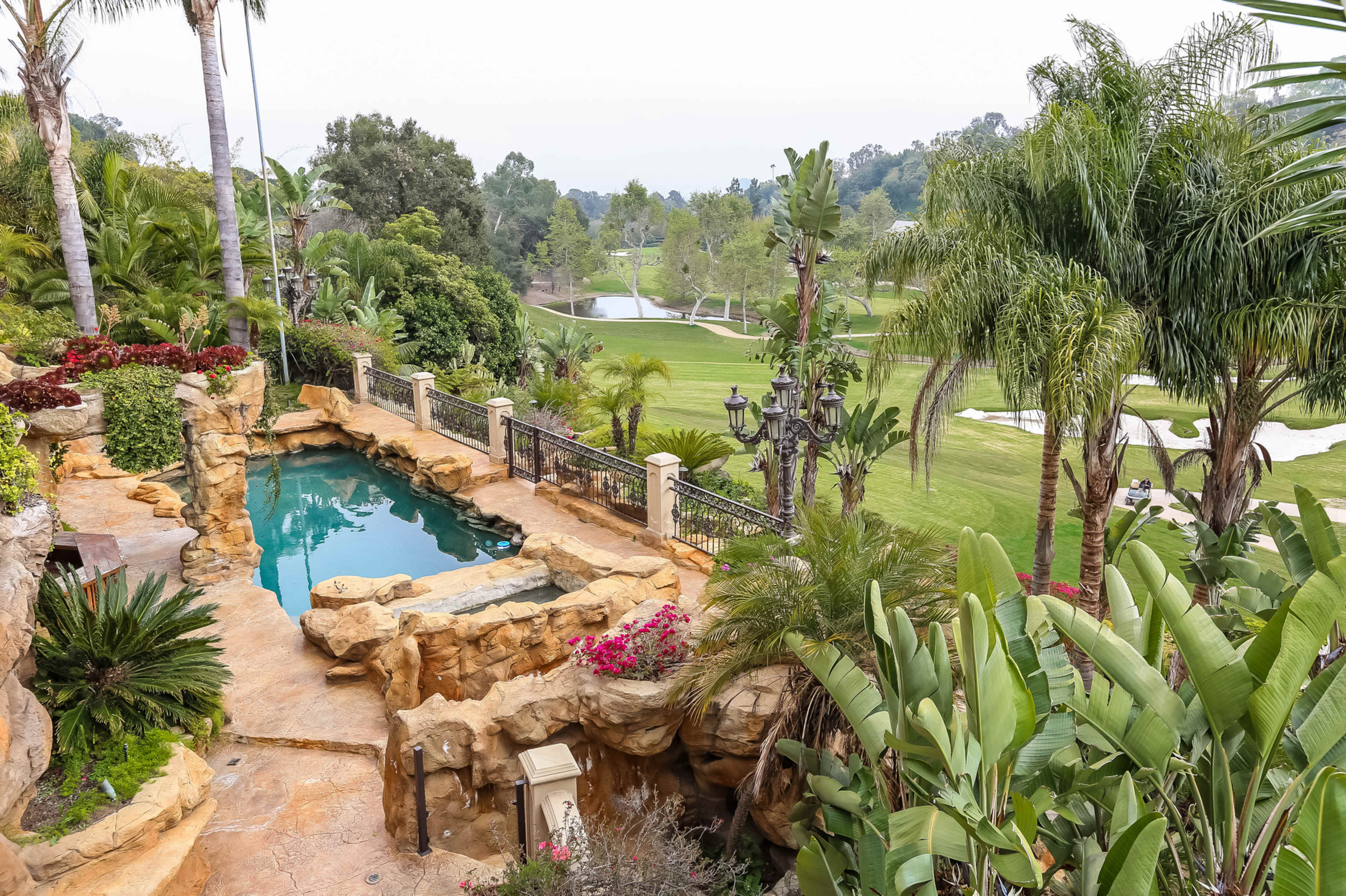 A luxurious backyard features a stone-accented swimming pool surrounded by lush greenery and palm trees, overlooking a golf course.