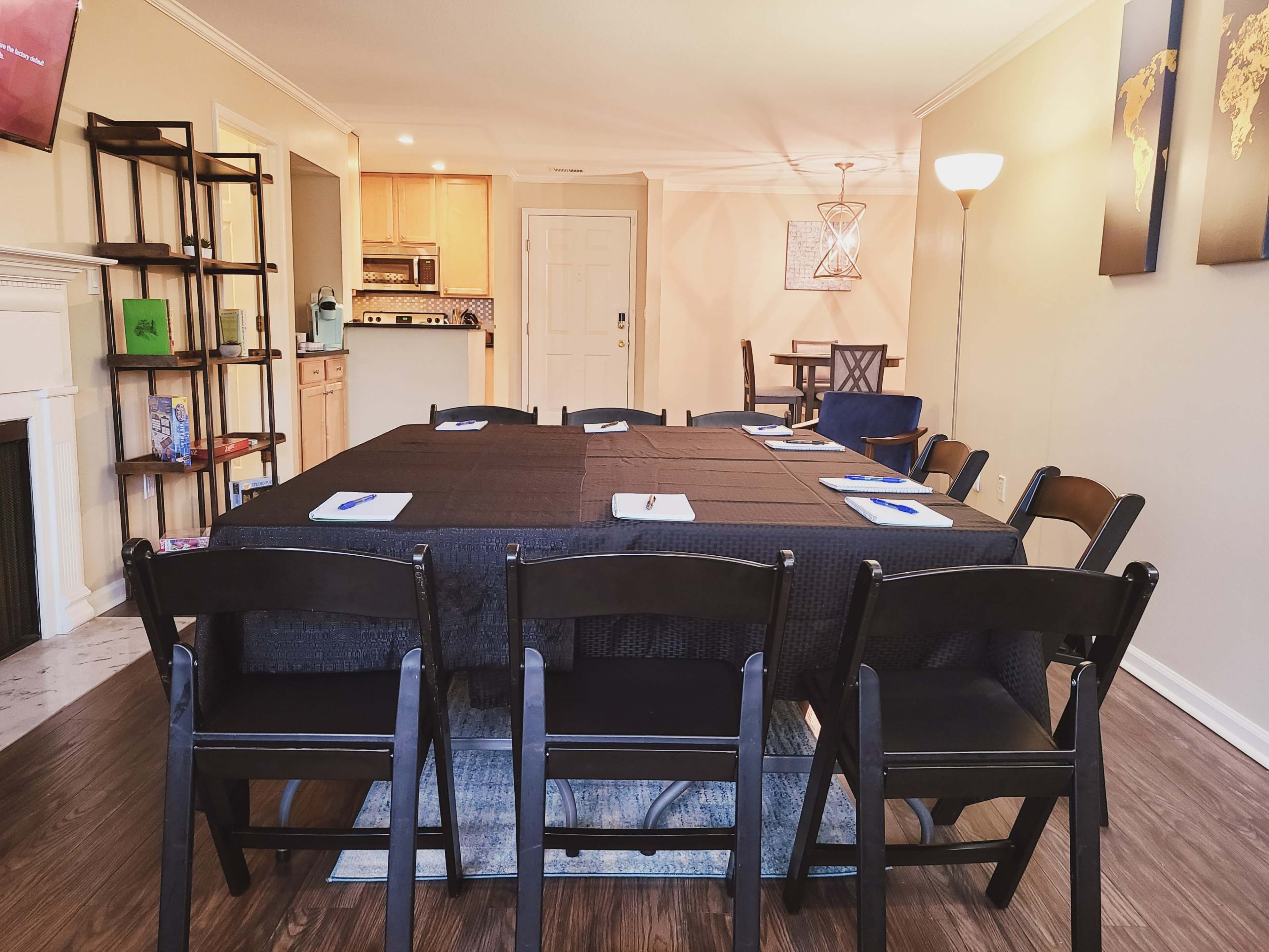 A long black table with chairs is set up in a dining area that connects to a kitchen and living space.