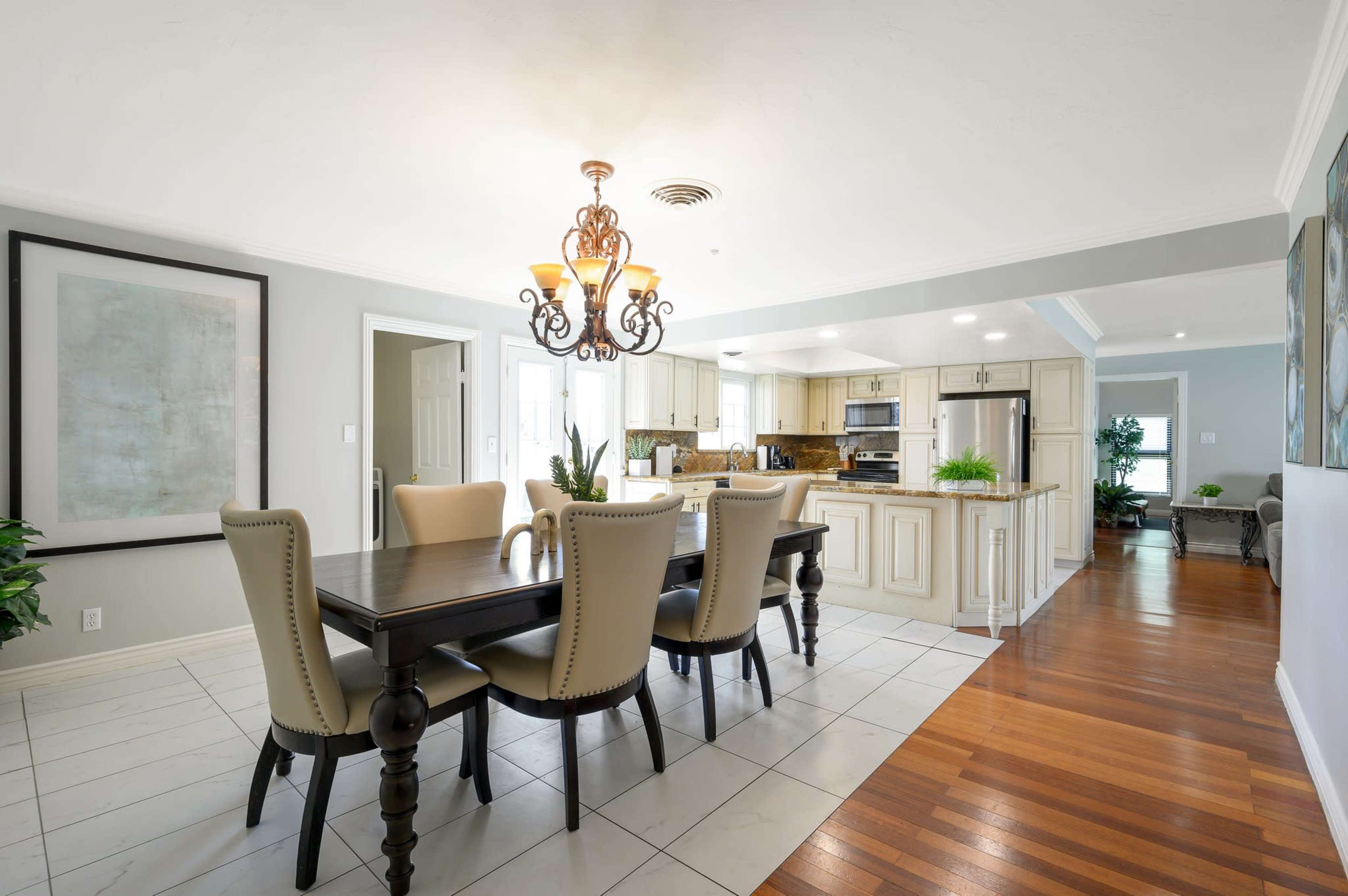 A dining area with a dark wooden table surrounded by beige chairs, illuminated by a chandelier, and adjacent to a kitchen with modern appliances.