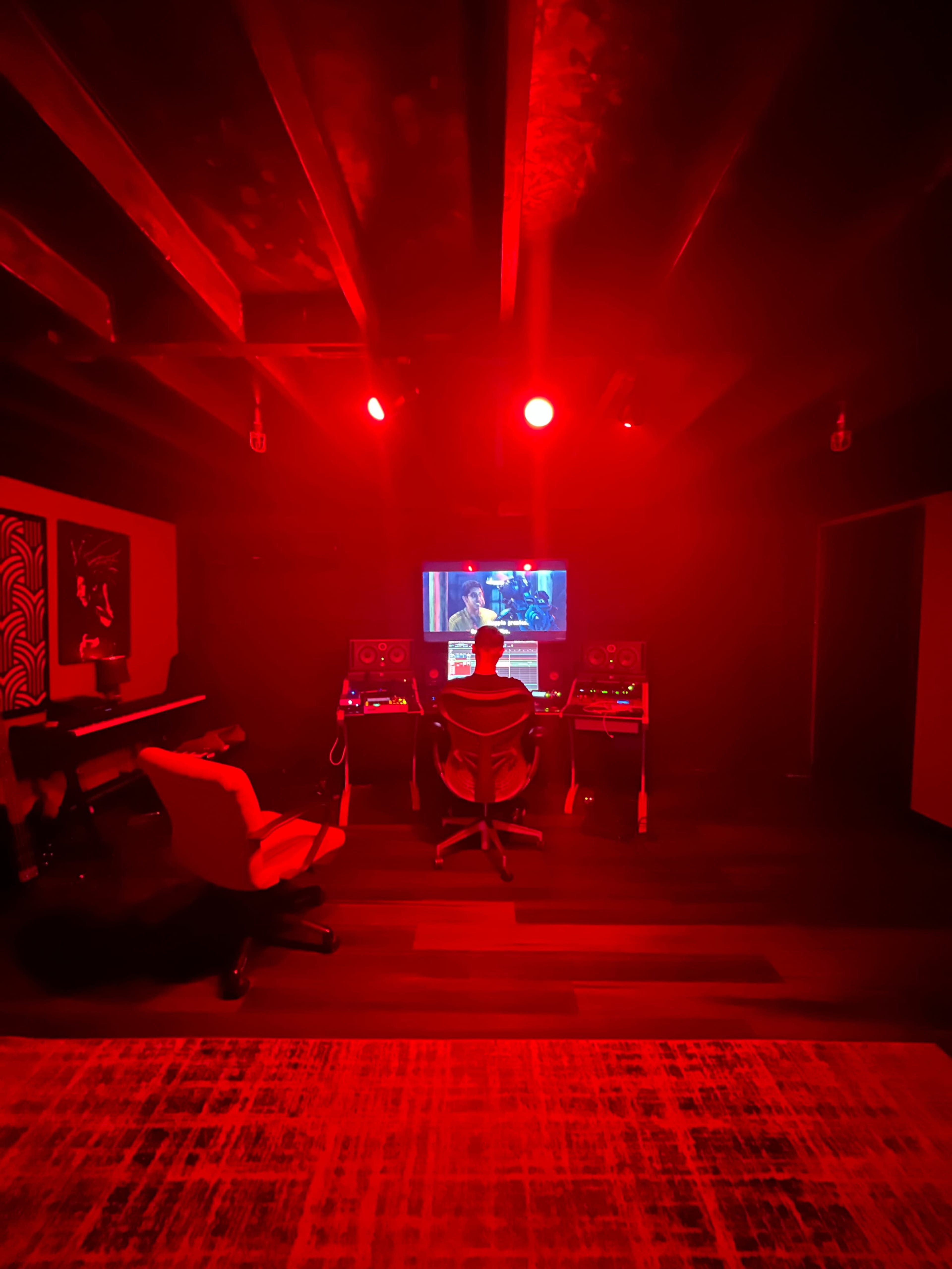 A person sits in a swivel chair in a dimly lit room, facing a computer screen with red lighting, surrounded by music equipment and a piano.
