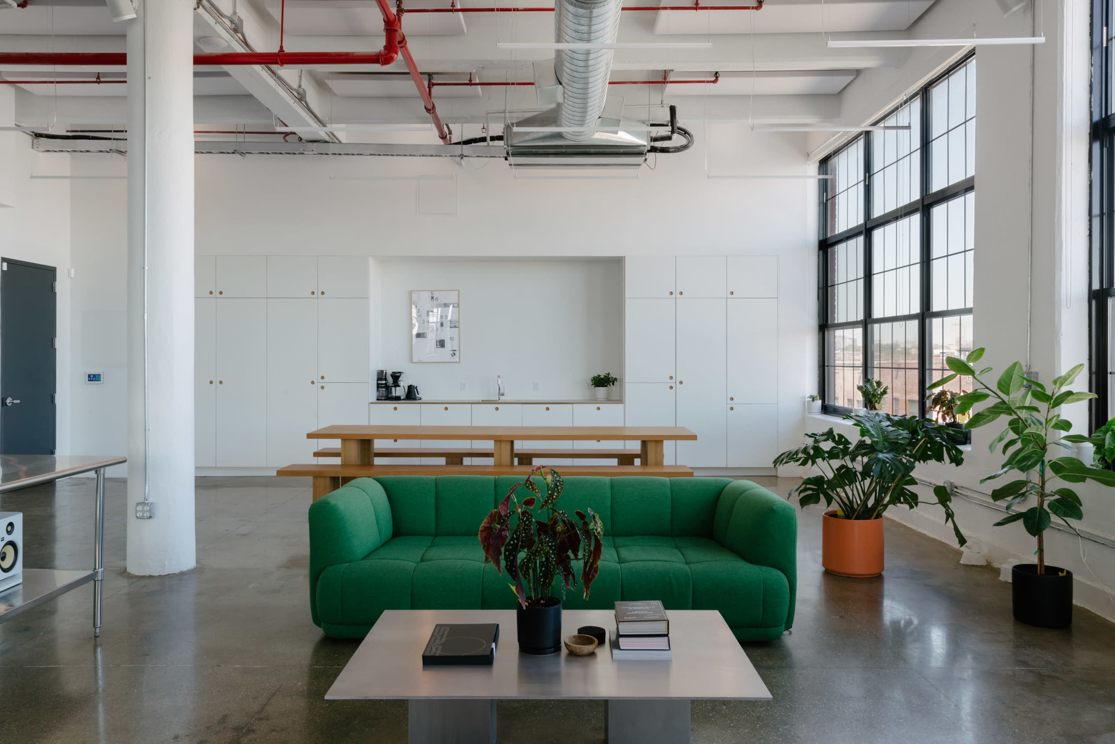 A modern, open-plan office space with a green sofa, a large table, and plants, featuring an expanse of windows that allow natural light to fill the area.