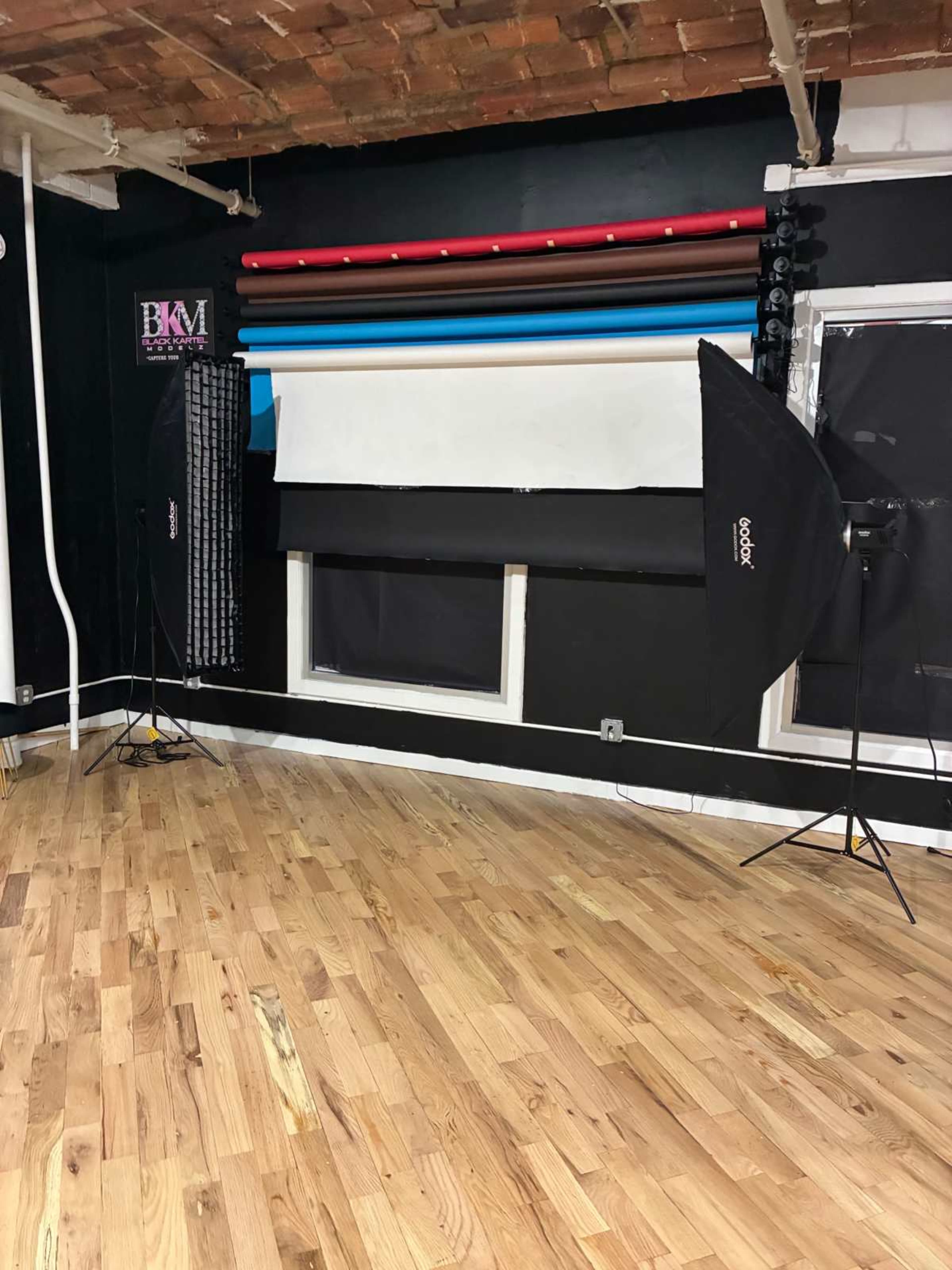The image shows a photography studio with wooden flooring, backdrop rolls in various colors on the wall, and two softbox lights positioned on either side of a window.