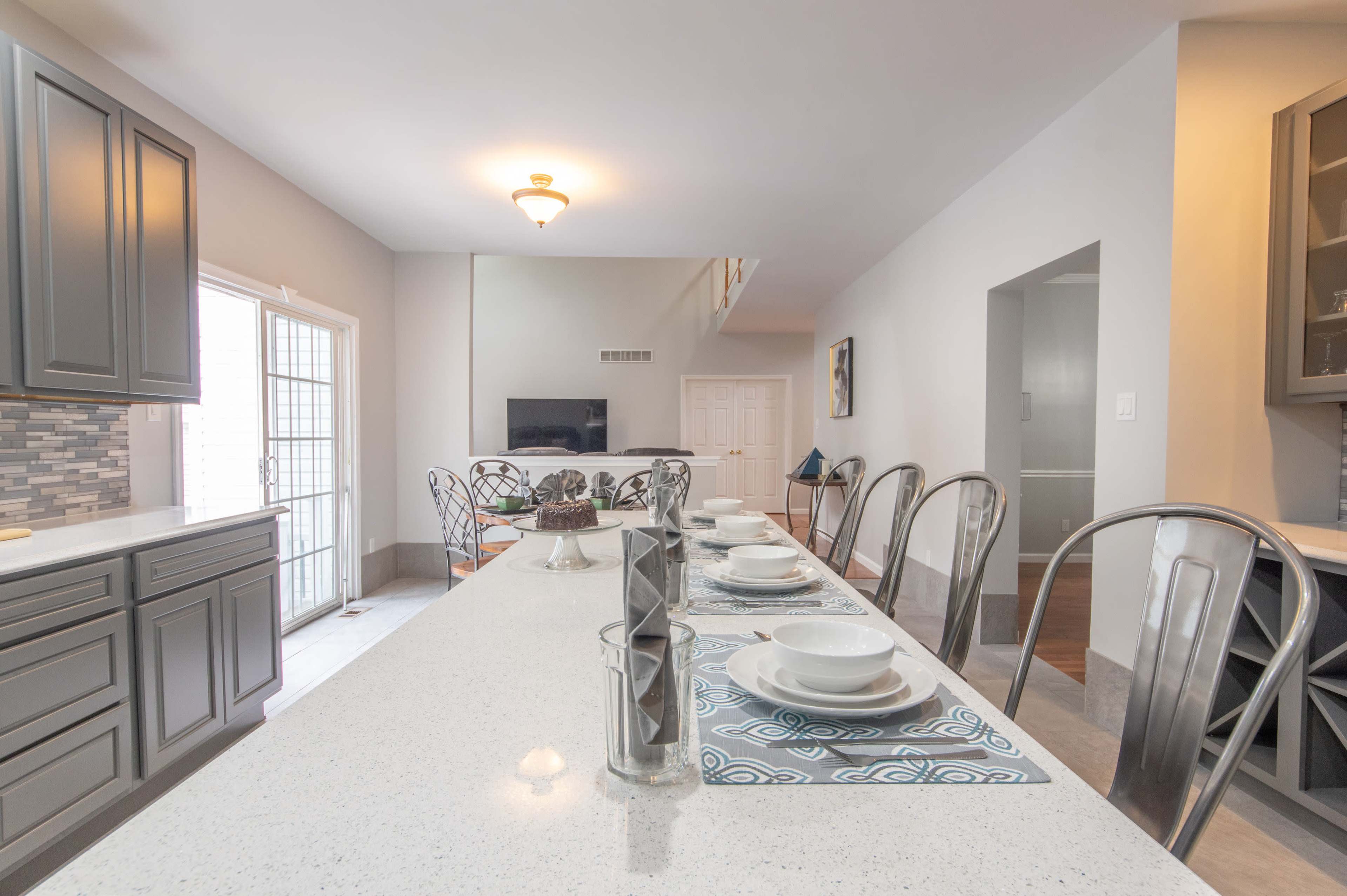 A spacious kitchen features a long countertop with set dining ware and a view of an adjoining living area with a television.