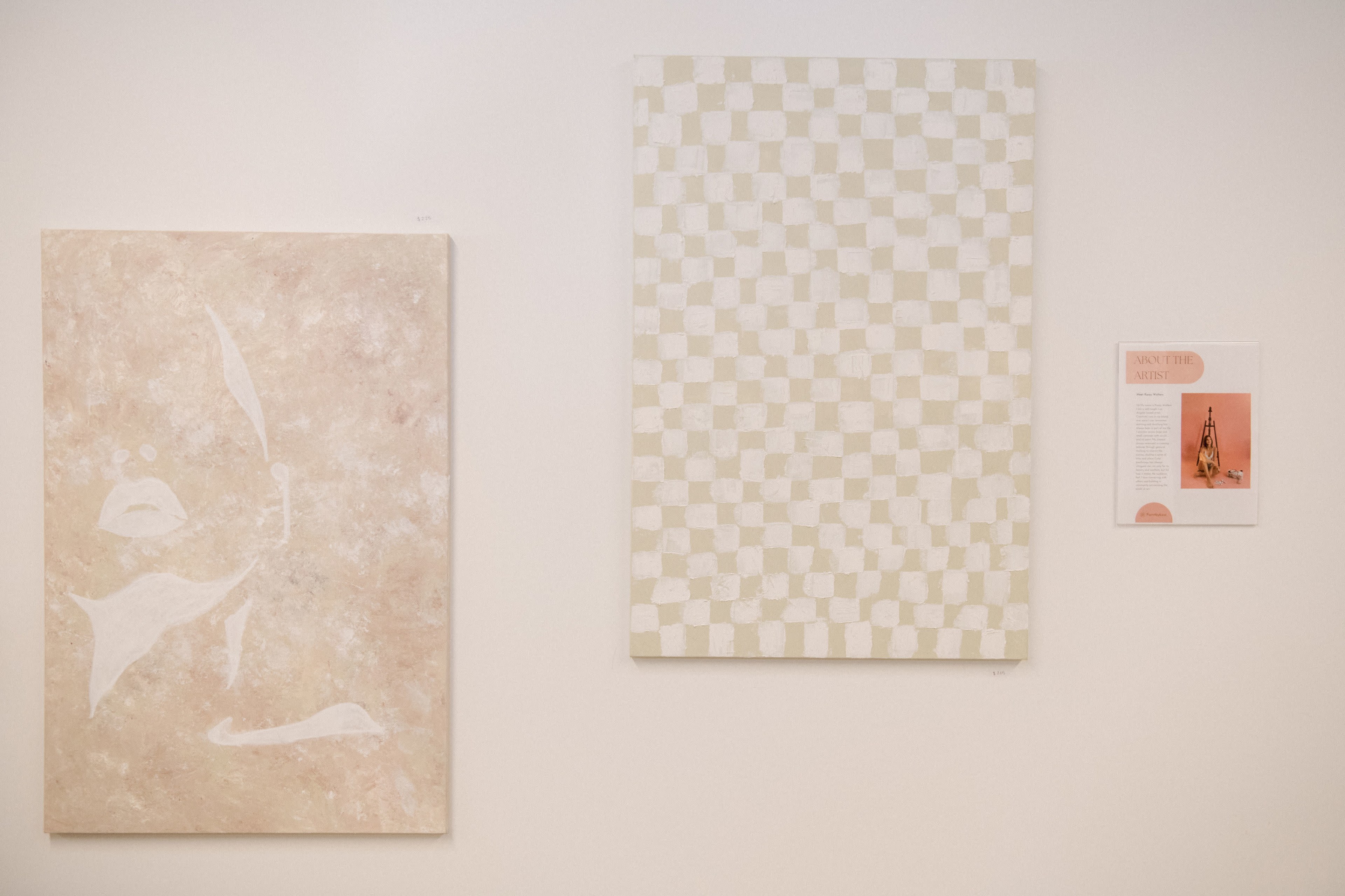 Two paintings hang on a white wall, with one featuring abstract forms in soft tones and the other displaying a grid pattern in muted colors, accompanied by an informational plaque.