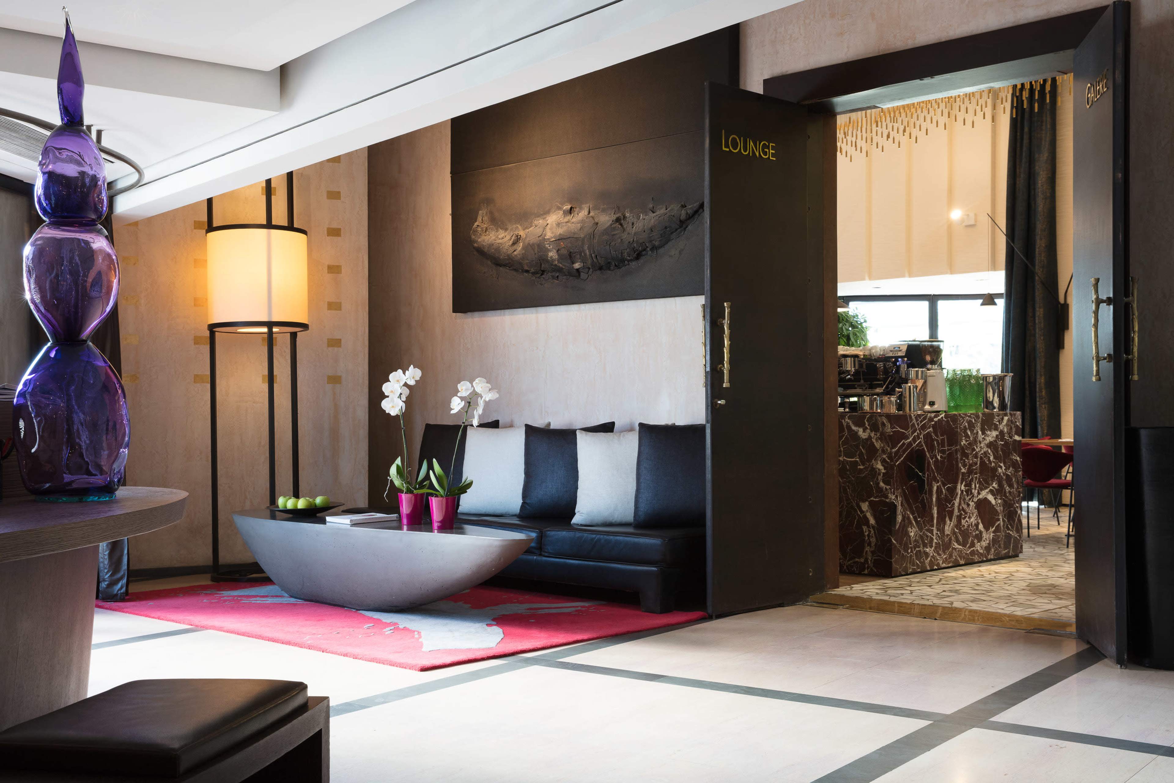 A modern hotel lobby features a lounge area with a large, minimalist coffee table, black seating, and decorative plants, alongside an entrance to a dining area.