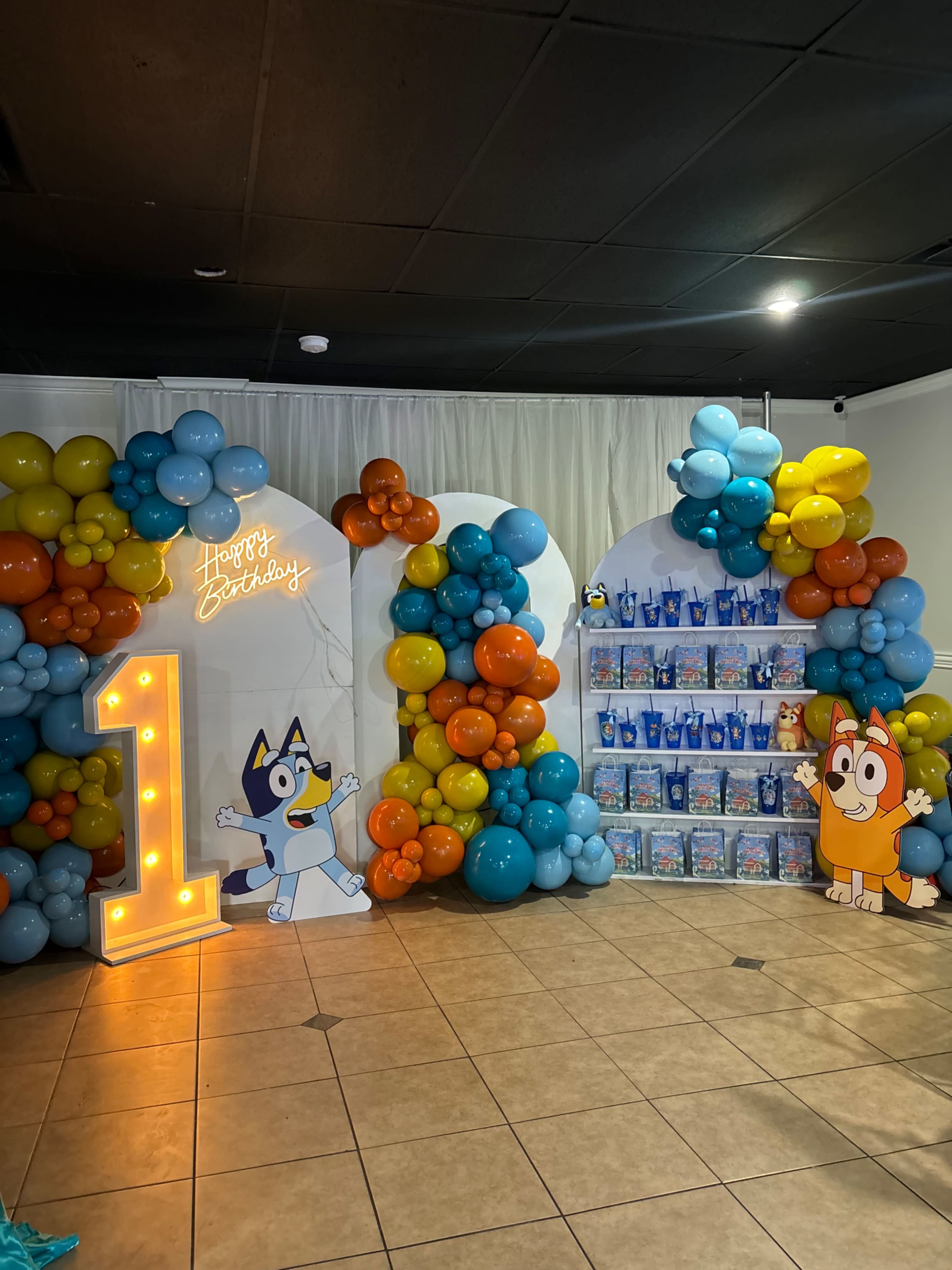 The scene features a decorated birthday party area with a large number "1," colorful balloons, and character displays from an animated show, along with gift bags on a shelf.