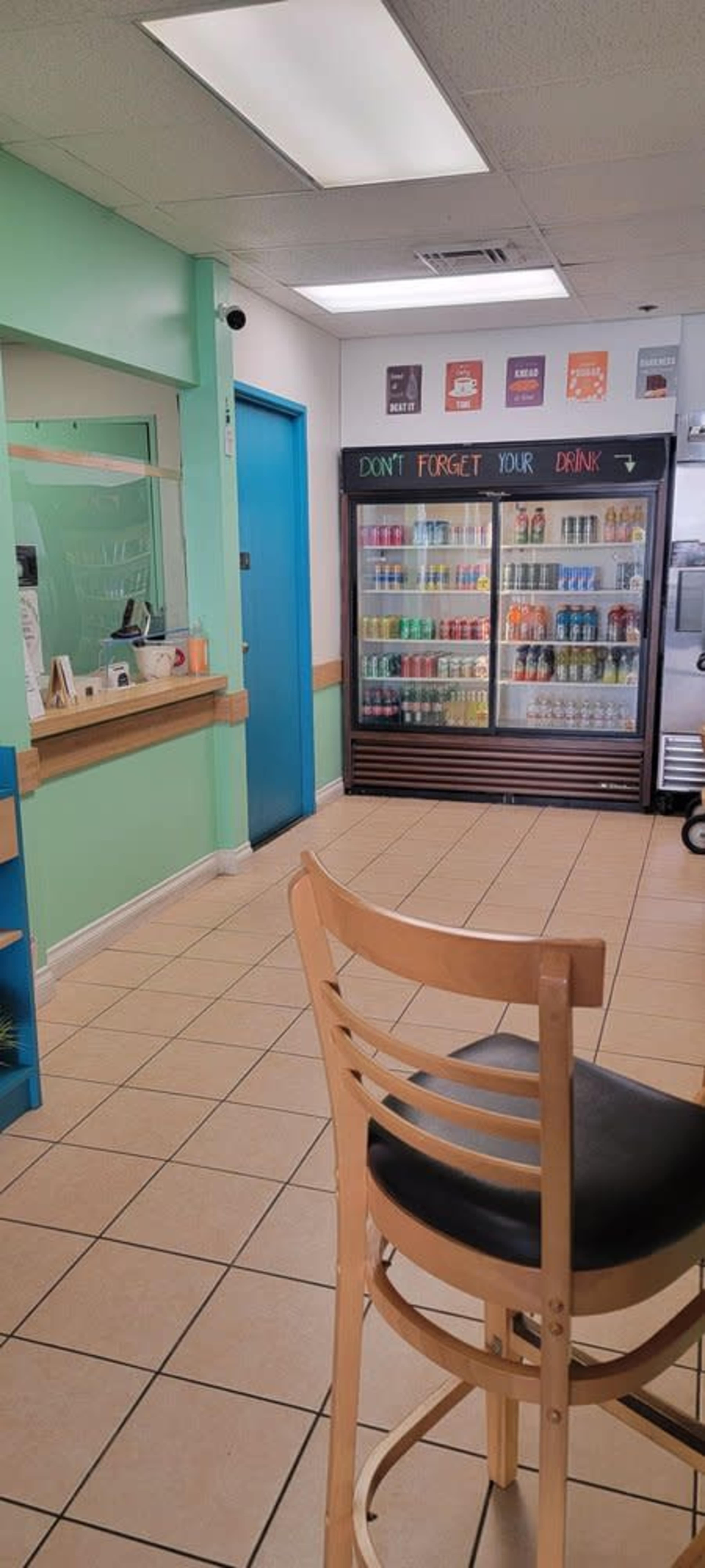 The image shows a small café or retail space with a counter, a large beverage refrigerator, and a bar stool positioned in the foreground.