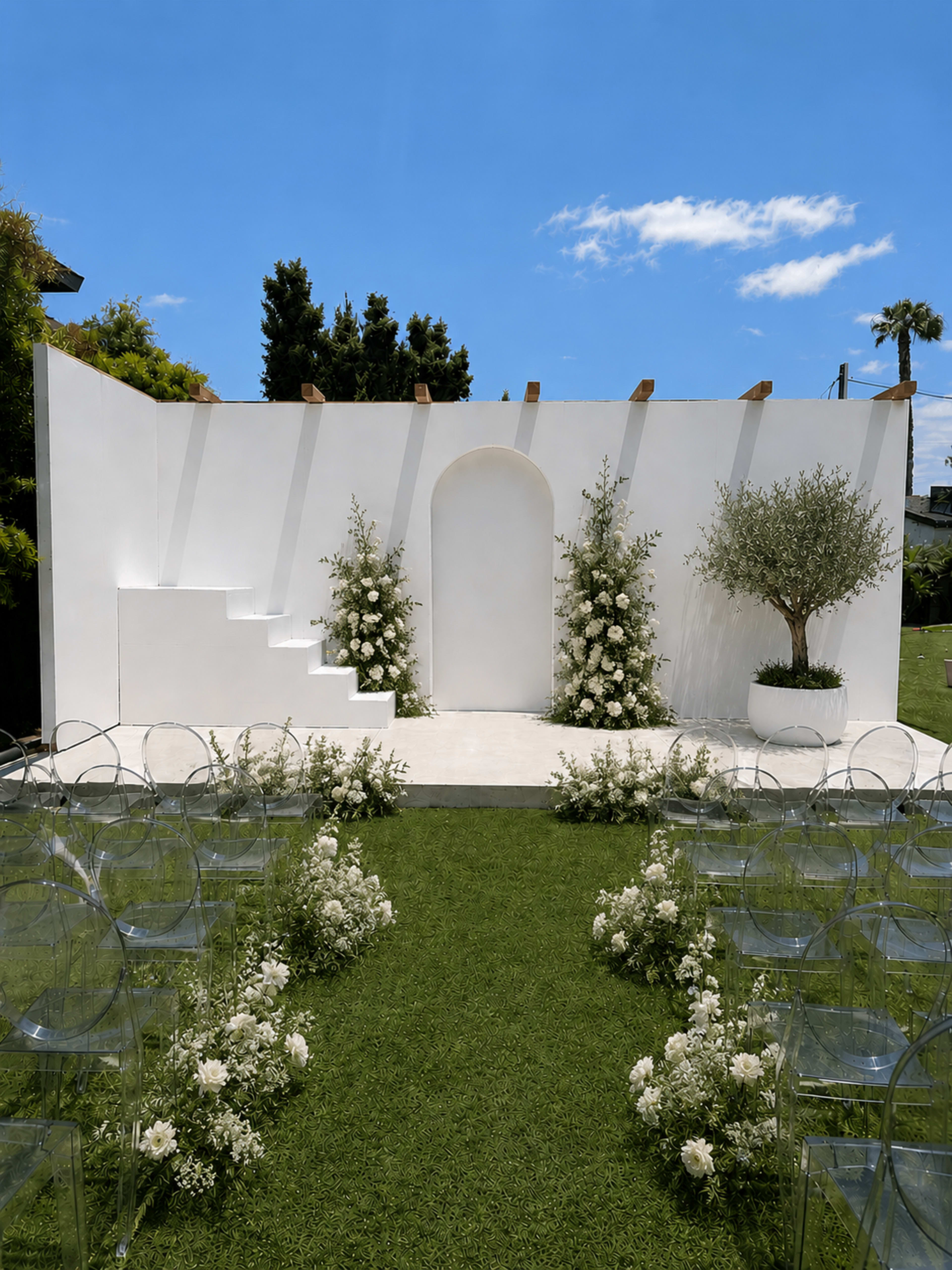 The image depicts a wedding ceremony setup featuring a minimalist white stage with floral arrangements, clear chairs aligned on either side, and a bright blue sky above.