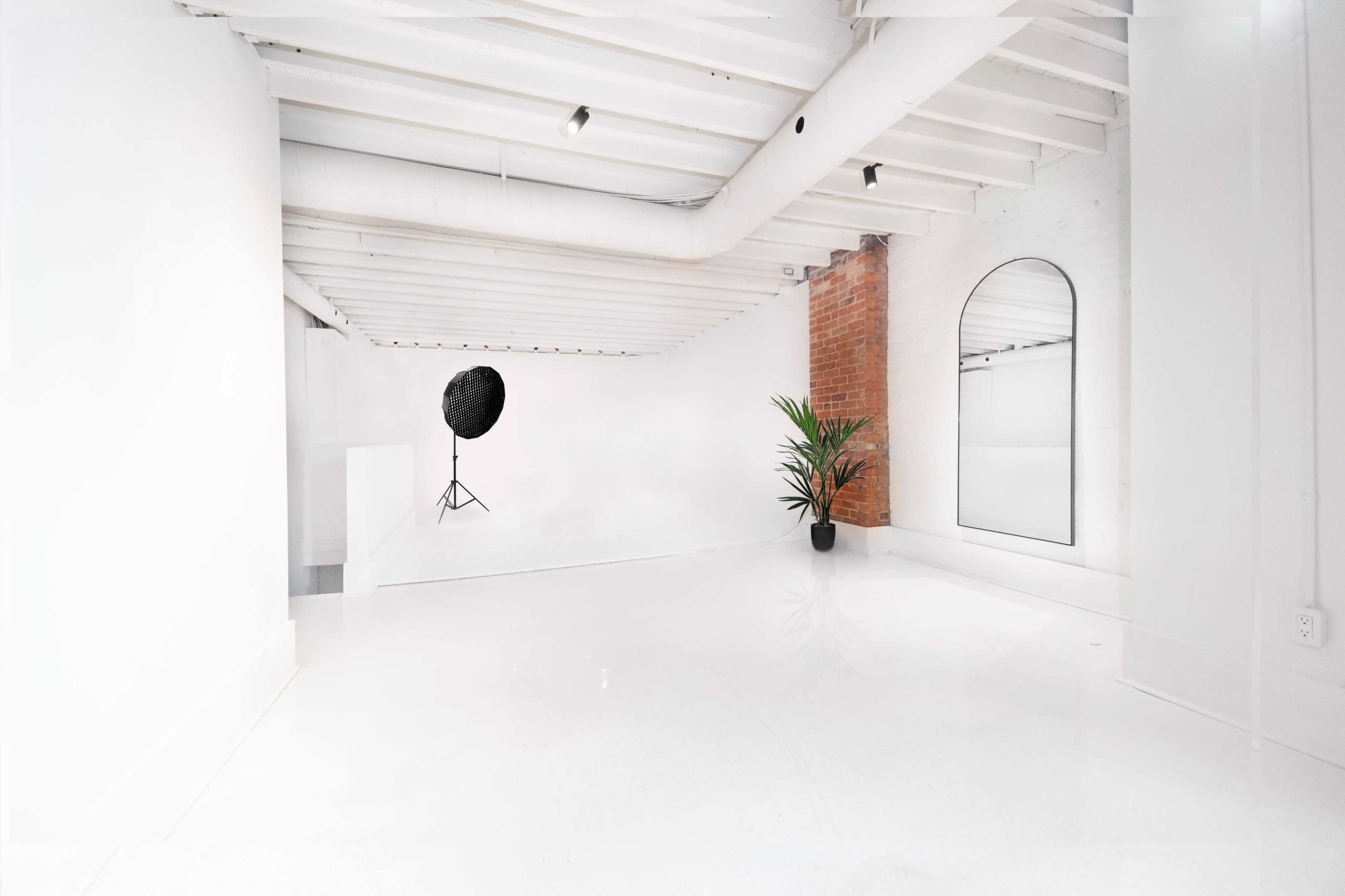 A minimalistic white room with a smooth floor, exposed beams, a large mirror, a black plant pot, and a lighting stand in the corner.