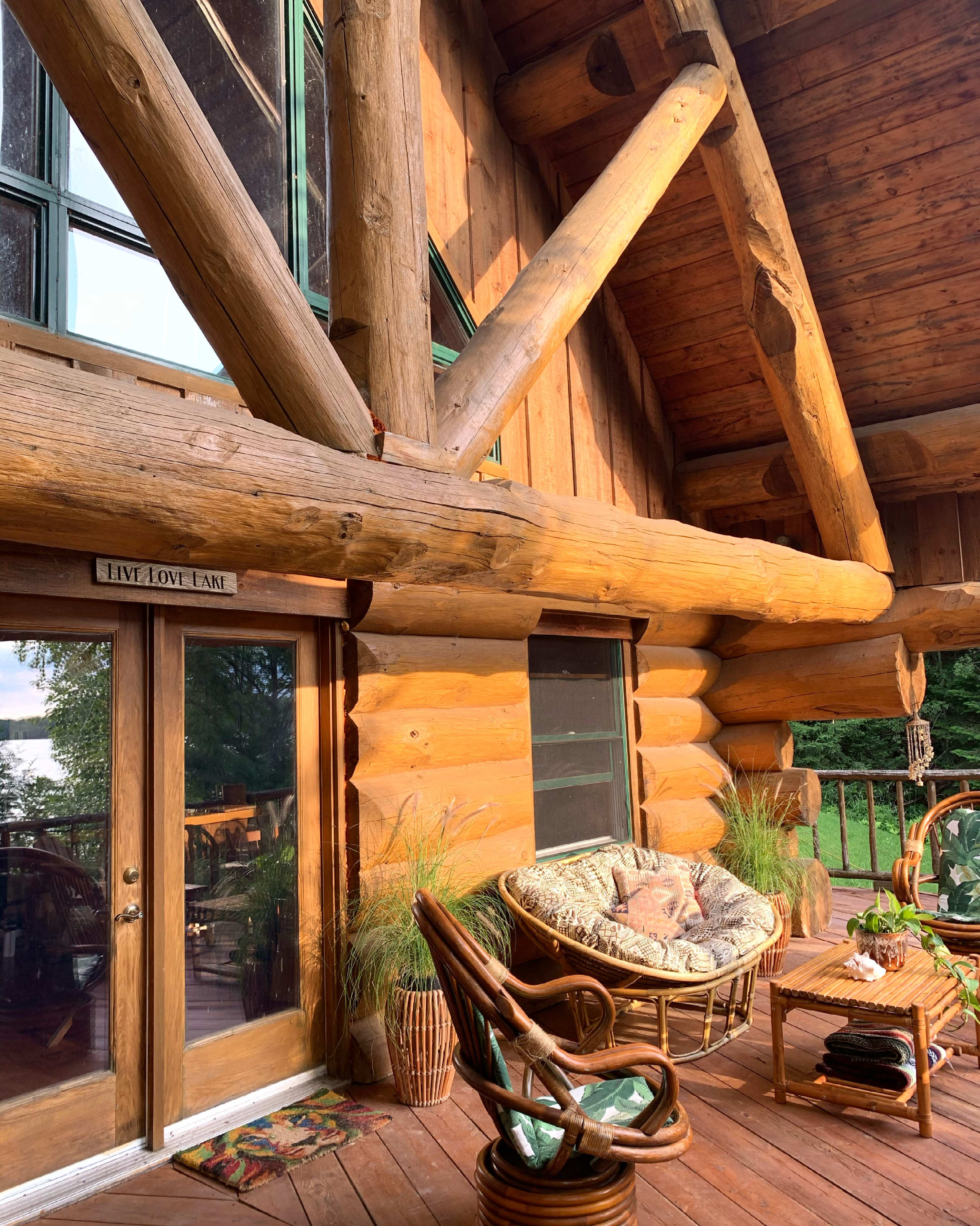 A rustic log cabin porch features a comfortable chair, potted plants, and large windows overlooking a serene landscape.