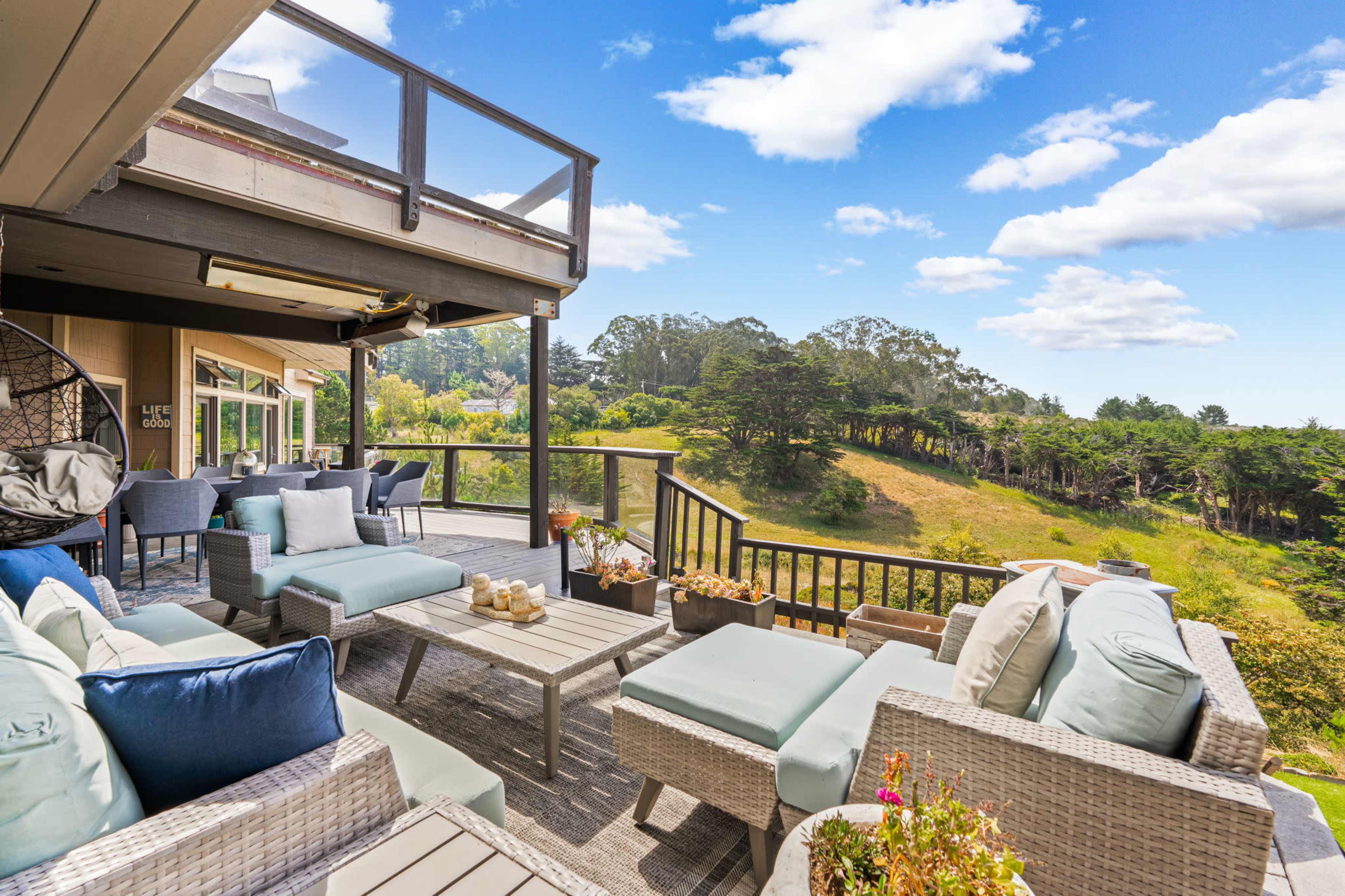 A spacious outdoor terrace features comfortable seating and a panoramic view of a green hillside under a blue sky with scattered clouds.