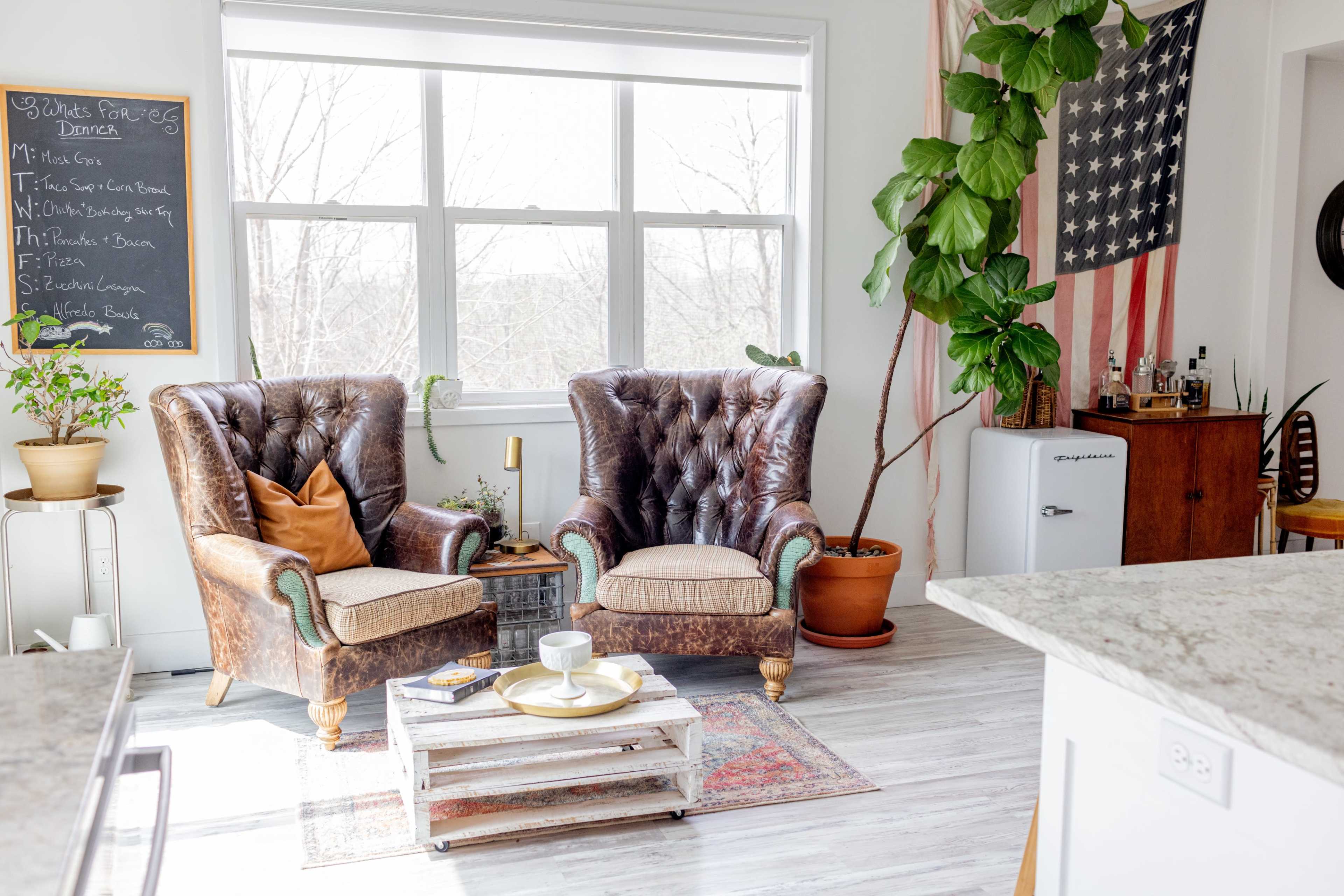 The image shows two vintage leather armchairs positioned next to a potted plant and an American flag hanging on the wall in a bright, airy room.